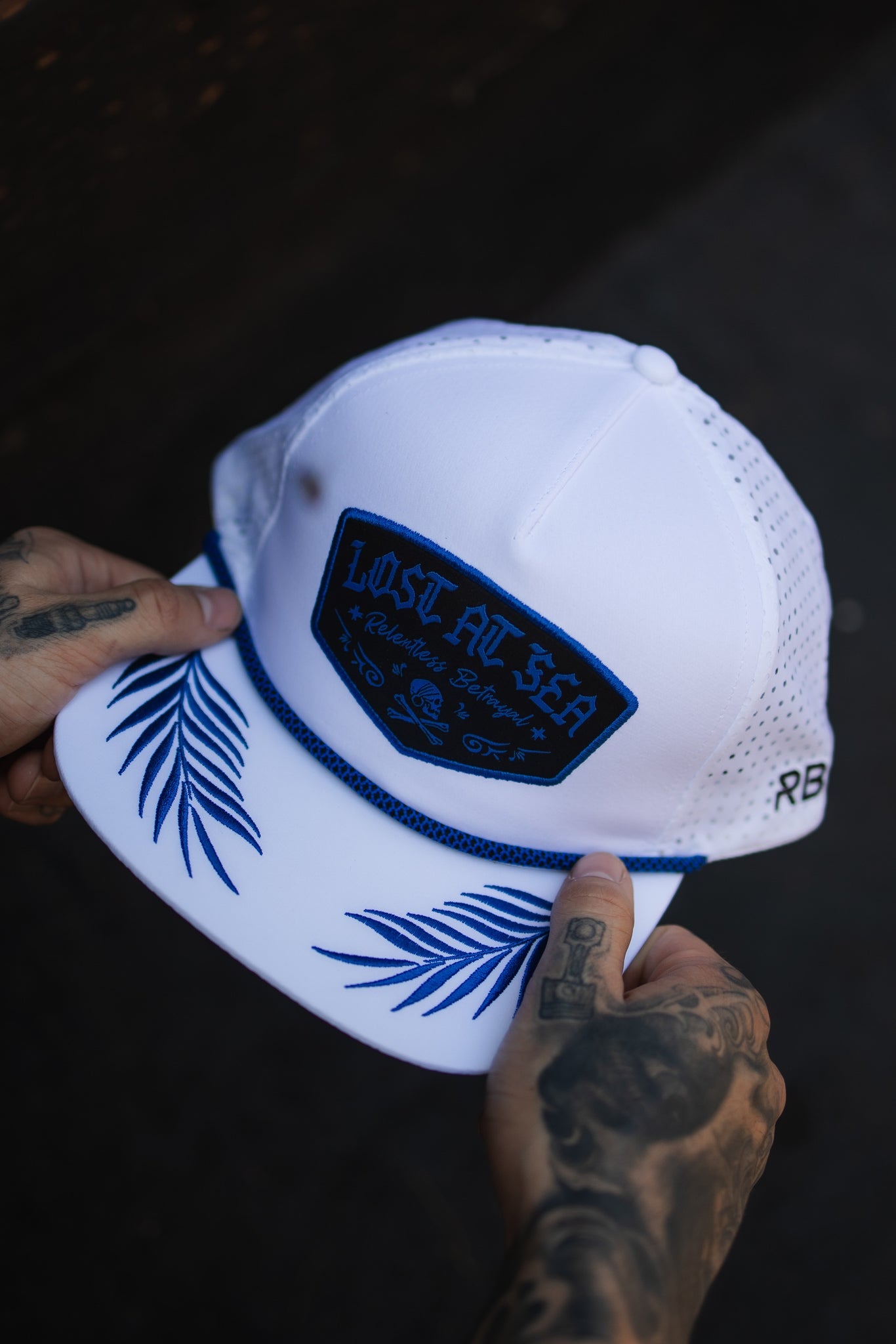 A tattooed individual holds the Lost at Sea Whiteout Snapback by Relentless Betrayal, a nautical white baseball cap with blue leaf patterns on the brim, a Lost at Sea patch, and a breathable perforated back panel.
