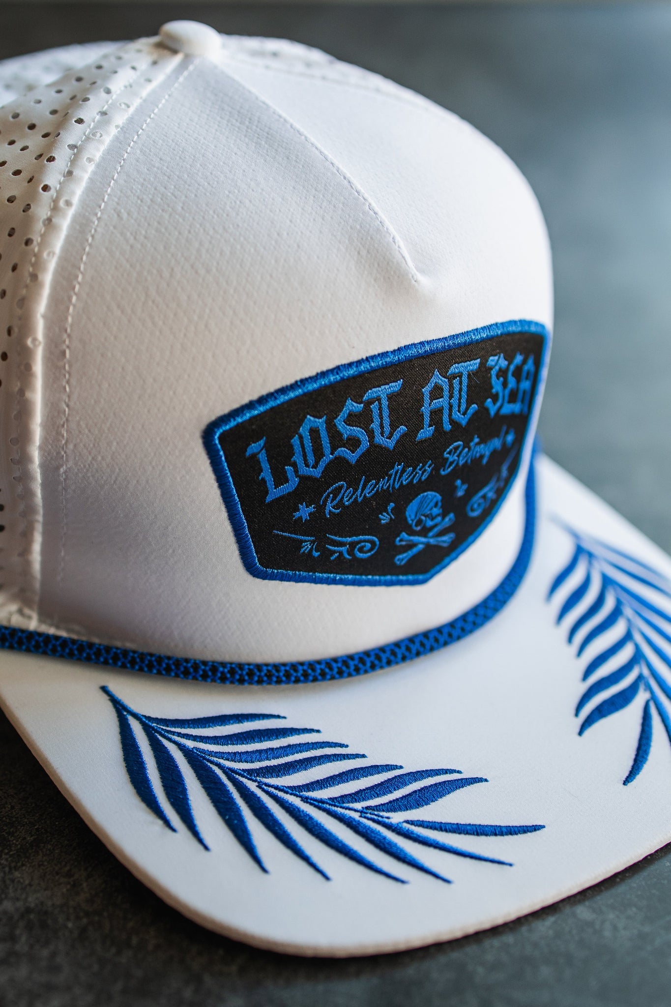 The Lost at Sea Whiteout Snapback by Relentless Betrayal is a white cap featuring a blue Lost At Sea patch and palm frond designs on the brim. Ventilation holes make it ideal for breezy days by the water.