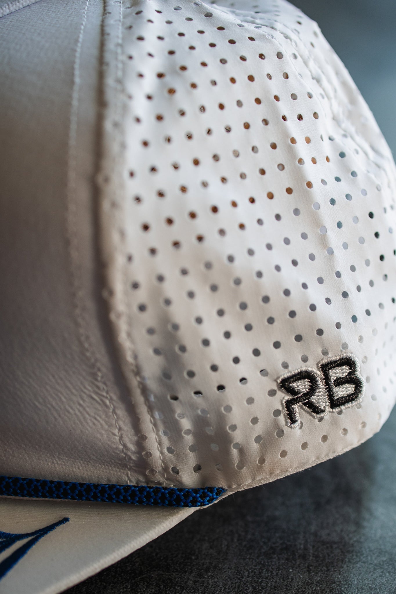 A close-up shows the Lost at Sea Whiteout Snapback by Relentless Betrayal, featuring a white perforated captain style with a blue brim. The initials RB are embroidered in black on the side, and small ventilation holes enhance its nautical charm.