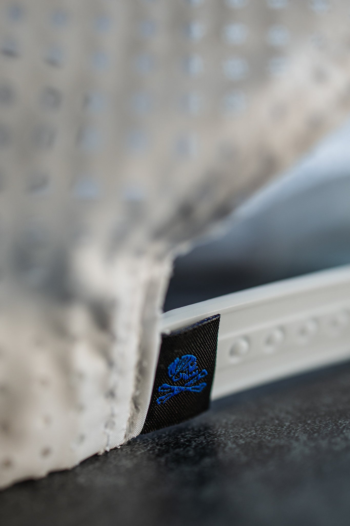 The image showcases the textures and details of the Lost at Sea Whiteout Snapback by Relentless Betrayal—a white perforated fabric and strap similar to a captains hat, with soft lighting highlighting its surface. A small black tag displays blue logo embroidery.