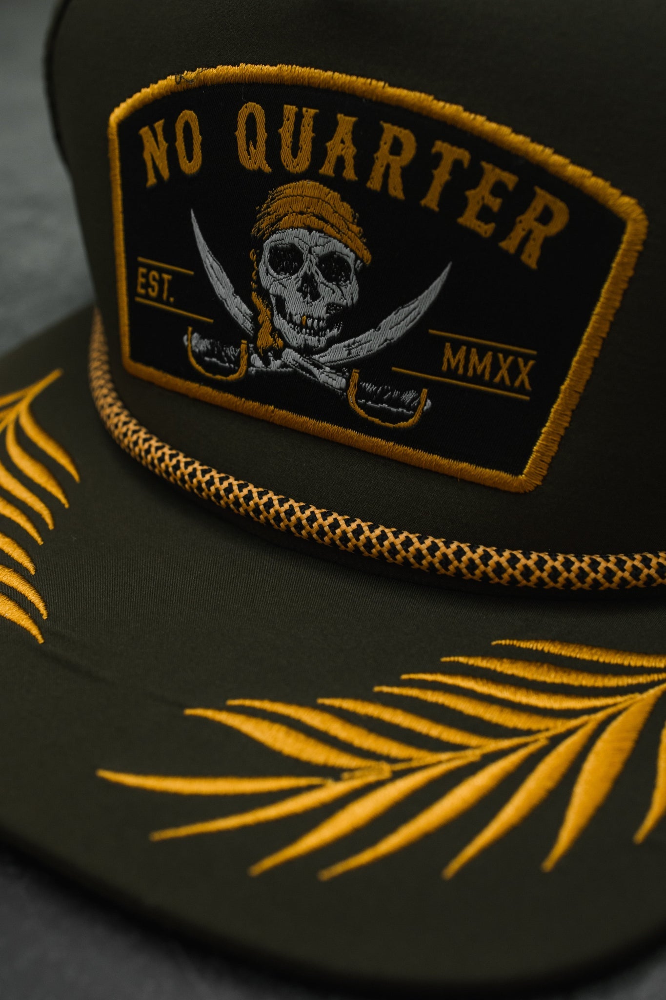 Close-up of Relentless Betrayals No Quarter Olive Snapback, showcasing yellow embroidery. The front panel features a skull with crossed swords beneath NO QUARTER and EST. MMXX, with palm leaf patterns elegantly on the brim.