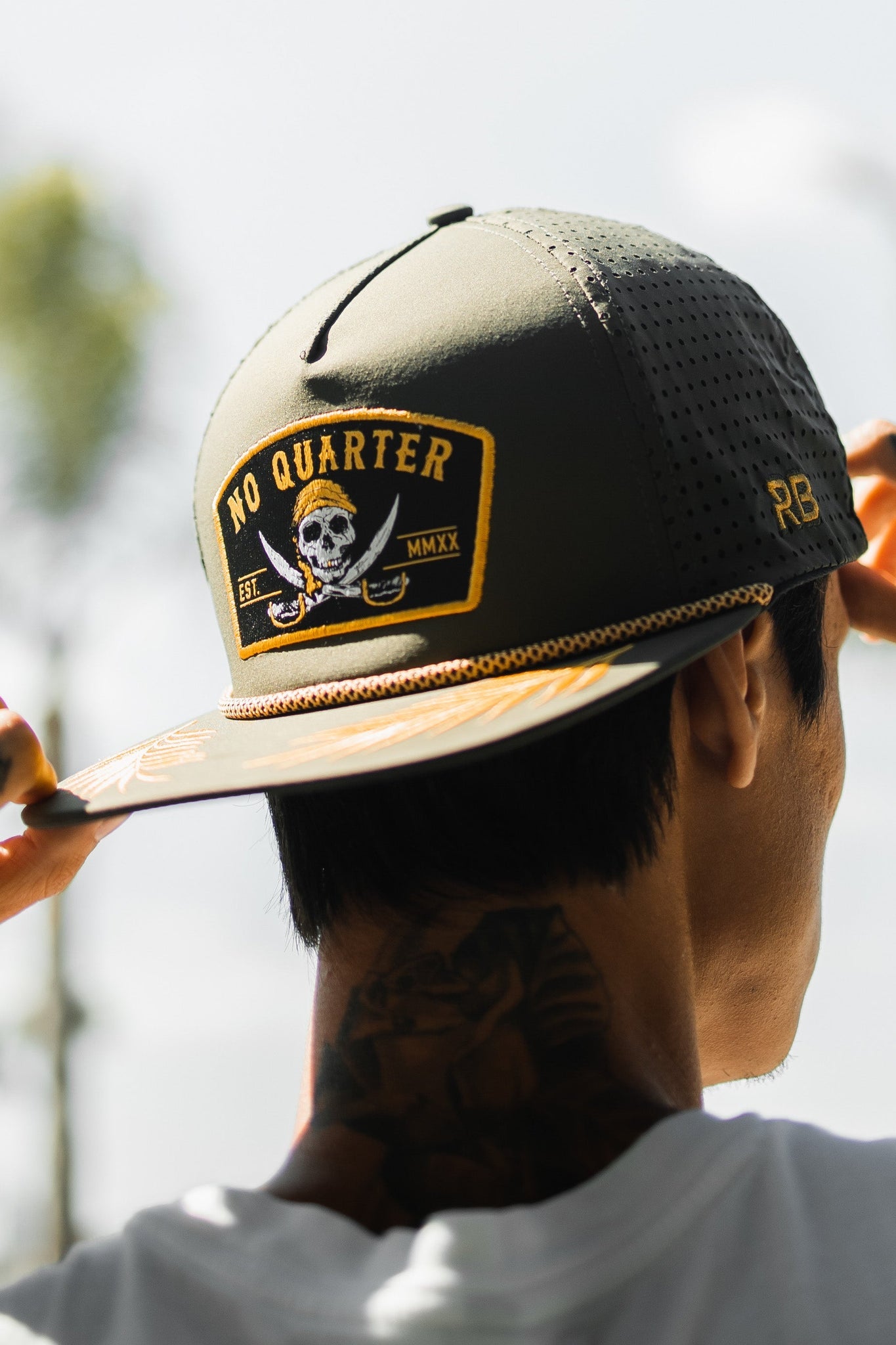 An individual wearing the No Quarter Olive Snapback by Relentless Betrayal, showcasing a skull and crossed swords patch, subtly adjusts the brim to reveal a neck tattoo. The softly blurred background enhances the mysterious allure of their style.
