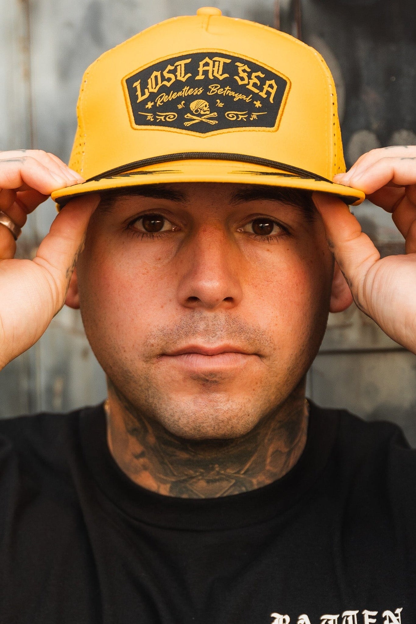 A person adjusts their Lost At Sea Gold Snapback by Relentless Betrayal, featuring vibrant yellow and a water-resistant design. Tattoos decorate their neck, complemented by a black t-shirt, while a blurred wall subtly frames the scene.