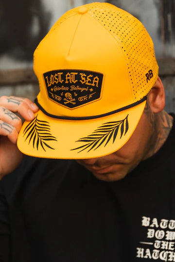 A person wearing the Relentless Betrayal Lost At Sea Gold Snapback features a skull design, casting a shadow over their face. Their tattooed fingers and black shirt with white text complement the snapbacks rugged charm.