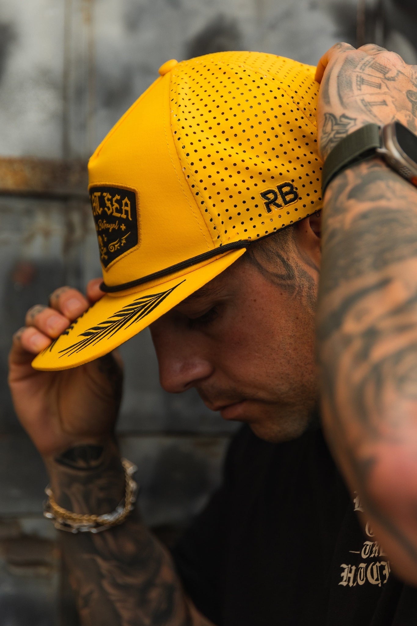 A person adjusts a Lost At Sea Gold Snapback from Relentless Betrayal, featuring black writing and patterns, while looking down. Tattoos cover their arms beneath a black shirt against a blurred neutral backdrop.