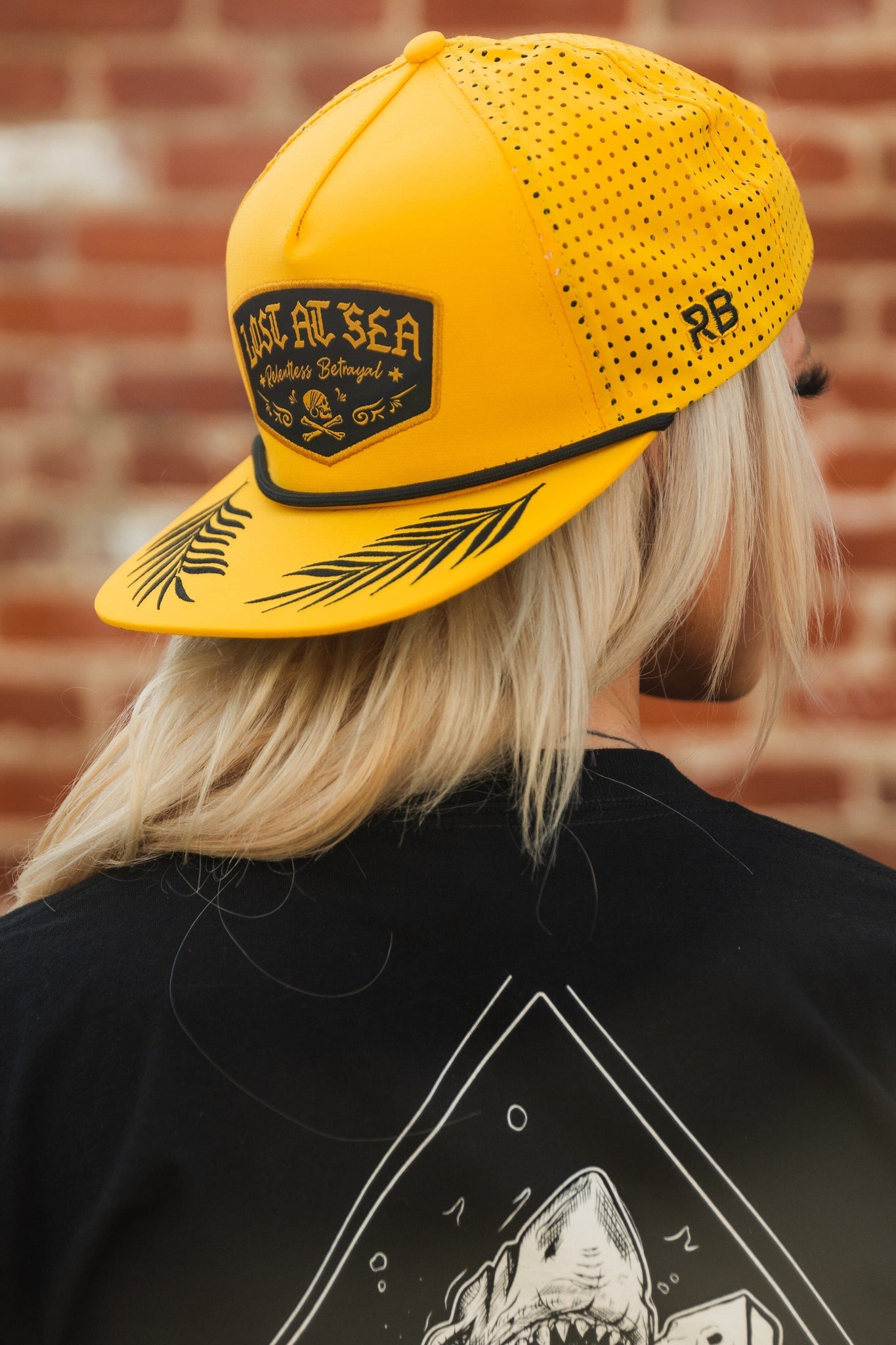 A person with long blonde hair wears the Lost At Sea Gold Snapback by Relentless Betrayal, featuring a snapback closure and patch reading LOST AT SEA, paired with a shark-themed black shirt against a brick wall.