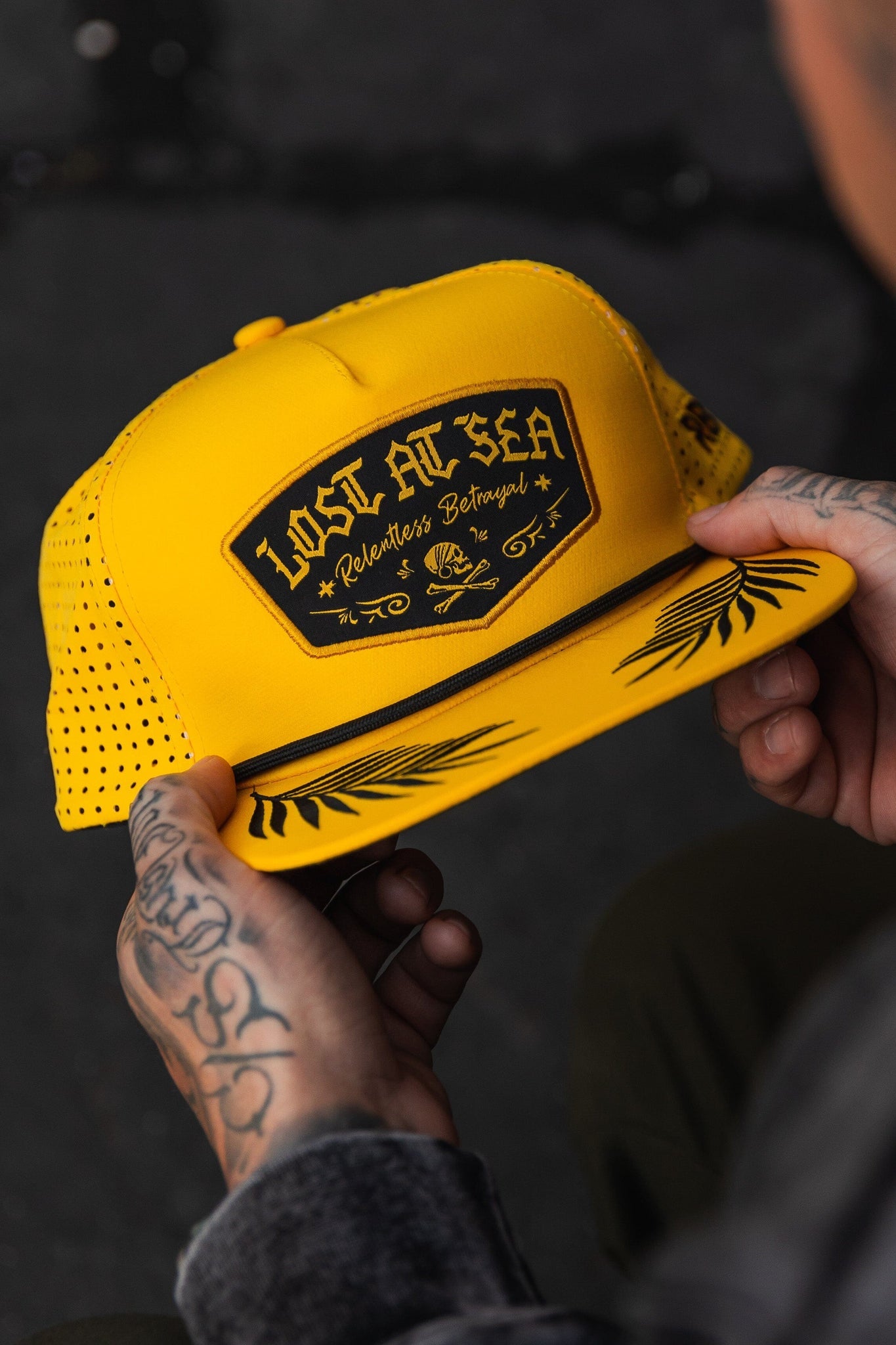 A person with tattooed hands holds the Lost At Sea Gold Snapback, a vibrant yellow cap by Relentless Betrayal. It features a black patch with skull design, water-resistant fabric, perforated sides, a palm leaf pattern on the brim, and an adjustable snapback enclosure.