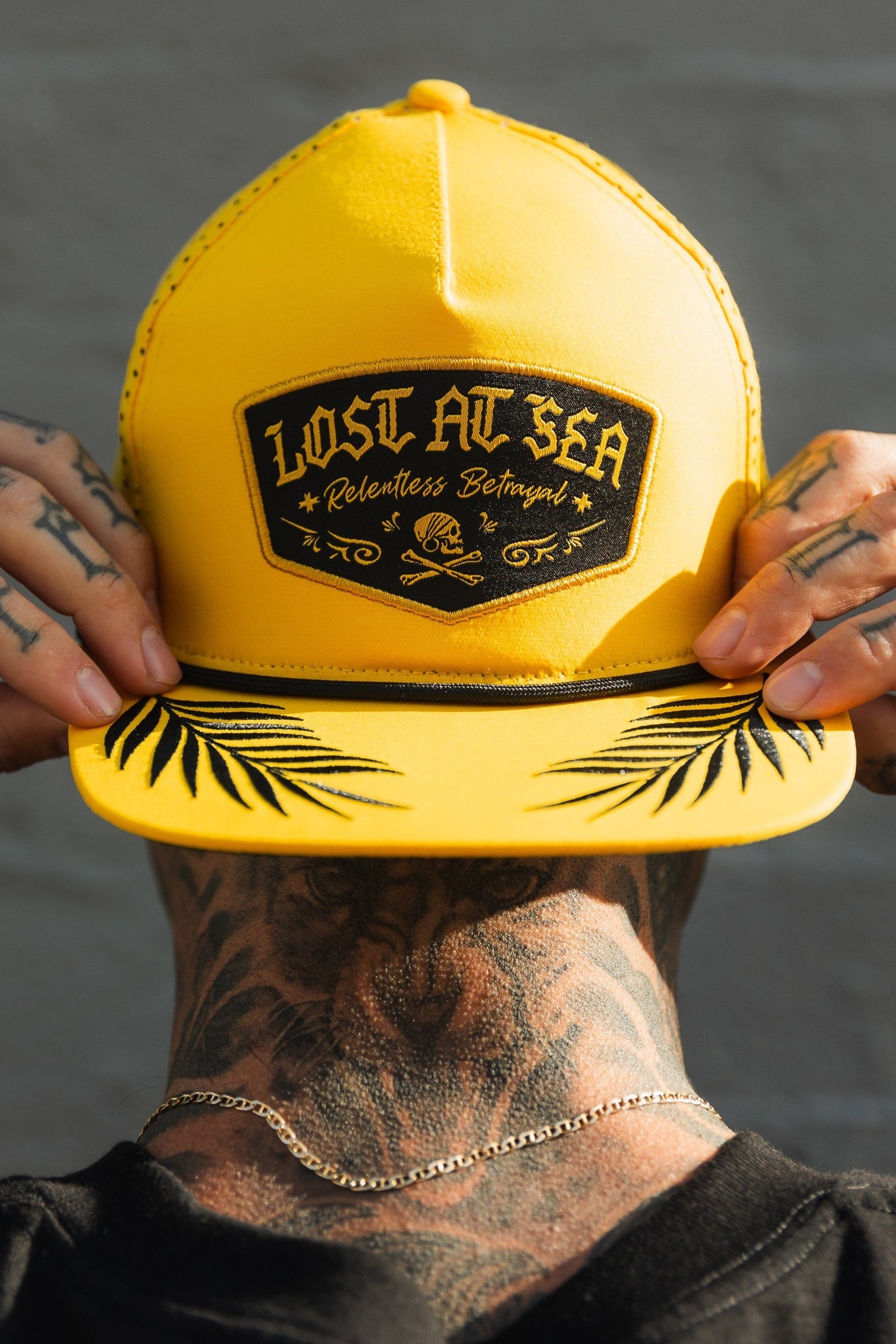 With tattooed hands and a neck tattoo, a person adjusts their Lost At Sea Gold Snapback from Relentless Betrayal, featuring a black patch with Lost at Sea and Relentless Betrayal, plus a skull and palm leaf design on its water-resistant brim.