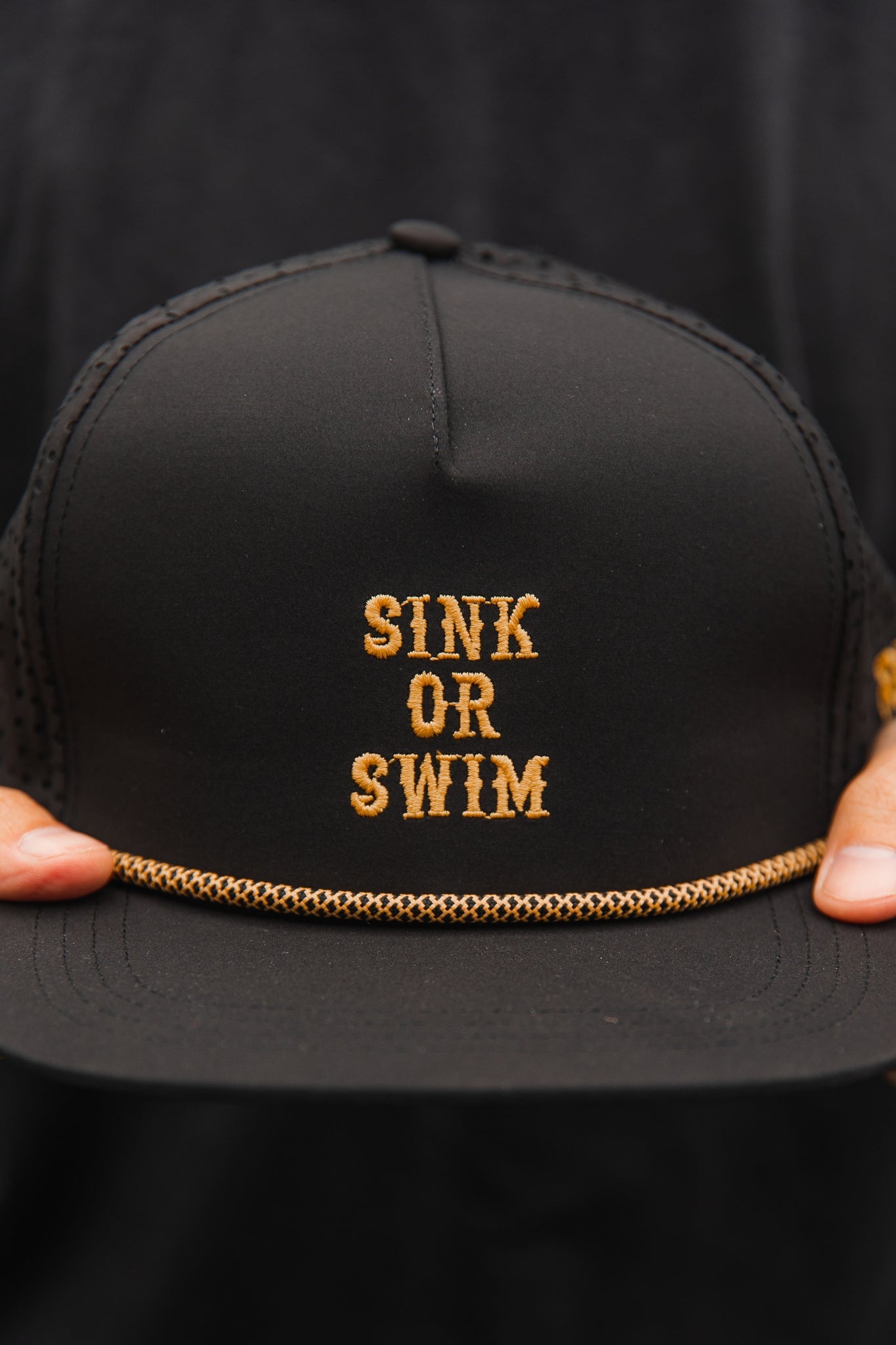 The Sink or Swim Blackout Snapback by Relentless Betrayal features gold embroidery with SINK OR SWIM on the front, a gold braided rope across the brim, and is elegantly displayed against a dark background.