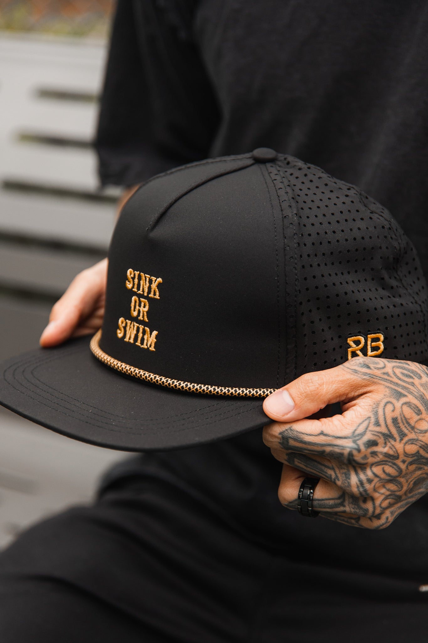 A person with tattooed hands holds a Sink or Swim Blackout Snapback by Relentless Betrayal, showcasing the embroidered SINK OR SWIM in gold, a gold chain above the brim, and side perforations for a sleek look.