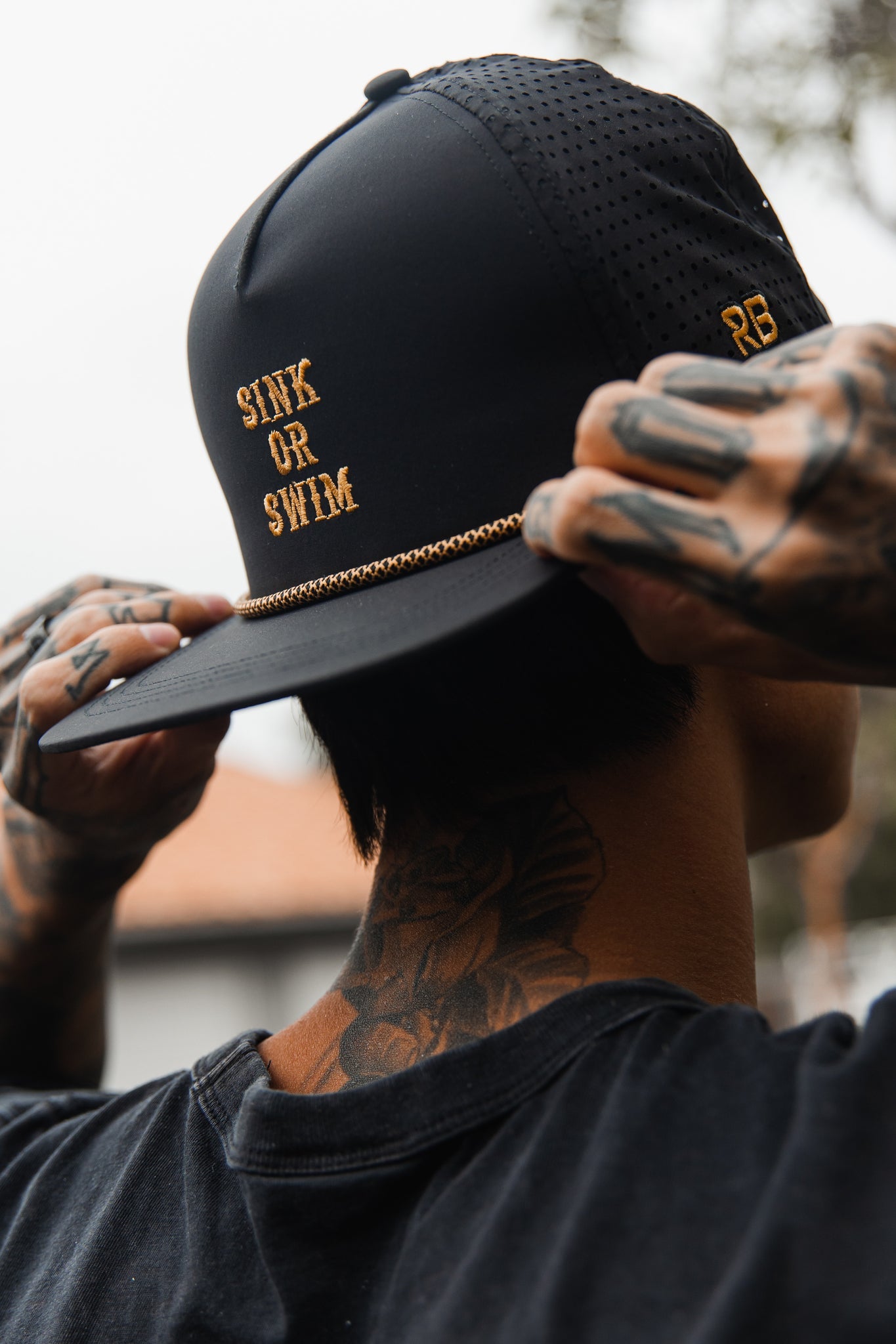 A person adjusts a Relentless Betrayal Sink or Swim Blackout Snapback with gold lettering and chain detail. Paired with tattooed hands, neck, and black shirt, they exude an effortlessly cool, edgy vibe.