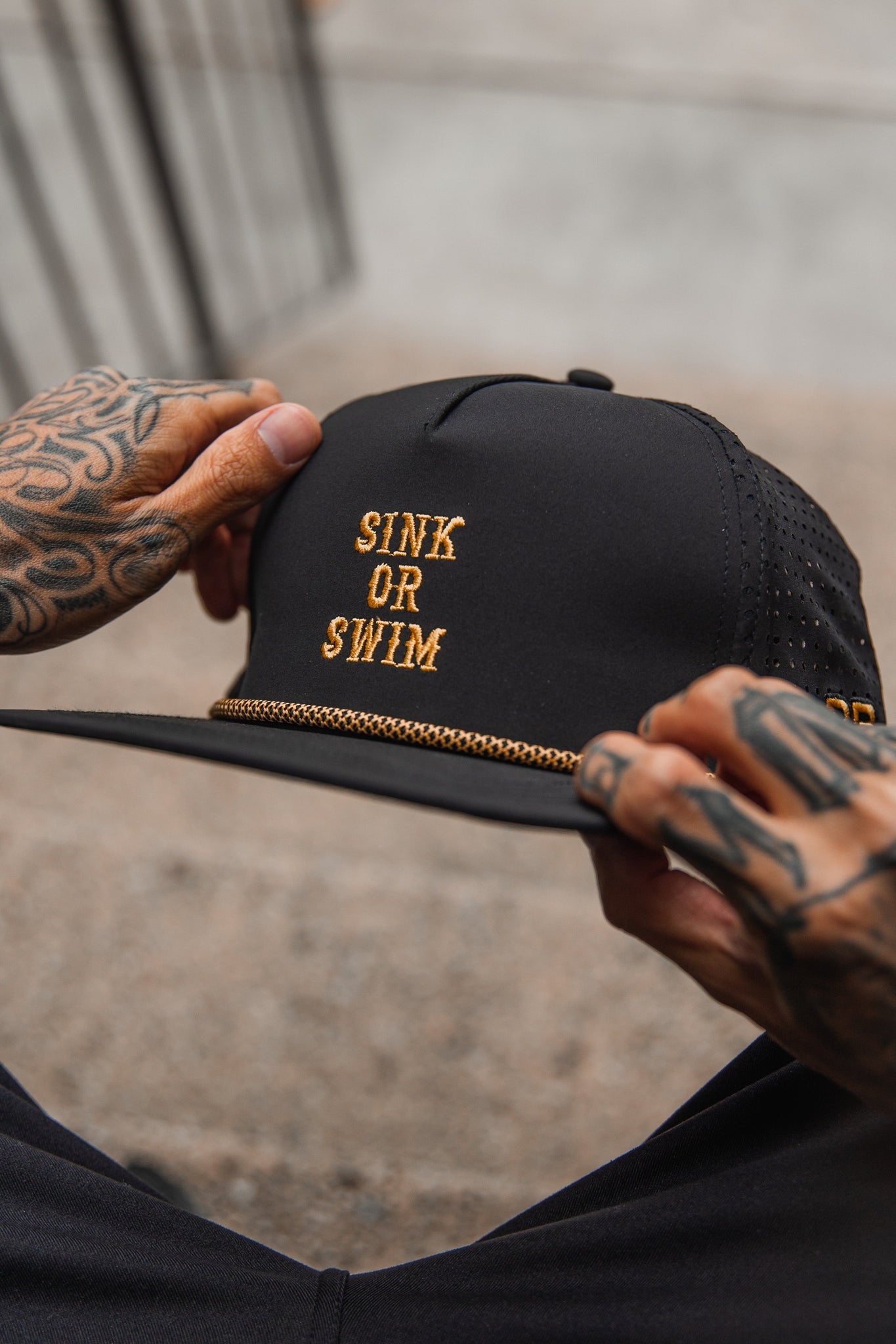 A person with tattooed hands holds the Relentless Betrayal Sink or Swim Blackout Snapback, featuring SINK OR SWIM in gold embroidery, a gold chain above the brim, and perforated fabric on the back. The backdrop is a blurred concrete surface.