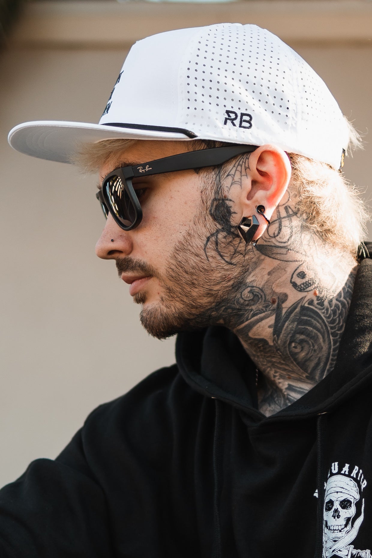 Person wearing a Sink or Swim WHITEOUT Snapback by Relentless Betrayal with ventilation holes, dark sunglasses, and a black hoodie with a skull design, showcasing neck tattoos and visible piercings. The cap combines style and comfort.