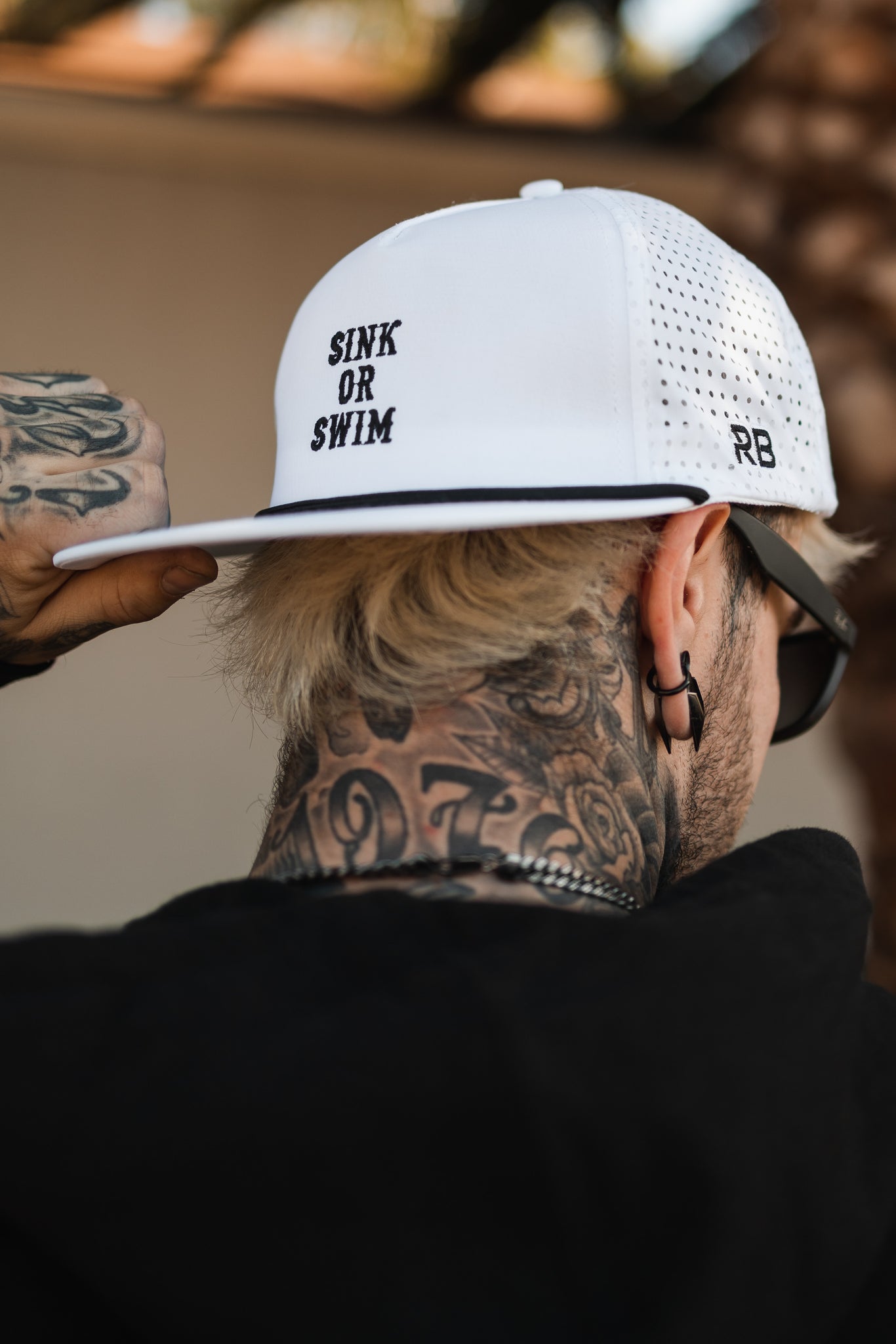 A blonde person with neck and hand tattoos adjusts their breathable Sink or Swim WHITEOUT Snapback by Relentless Betrayal. The white cap, embroidered with SINK OR SWIM, complements their black sunglasses and dark top.