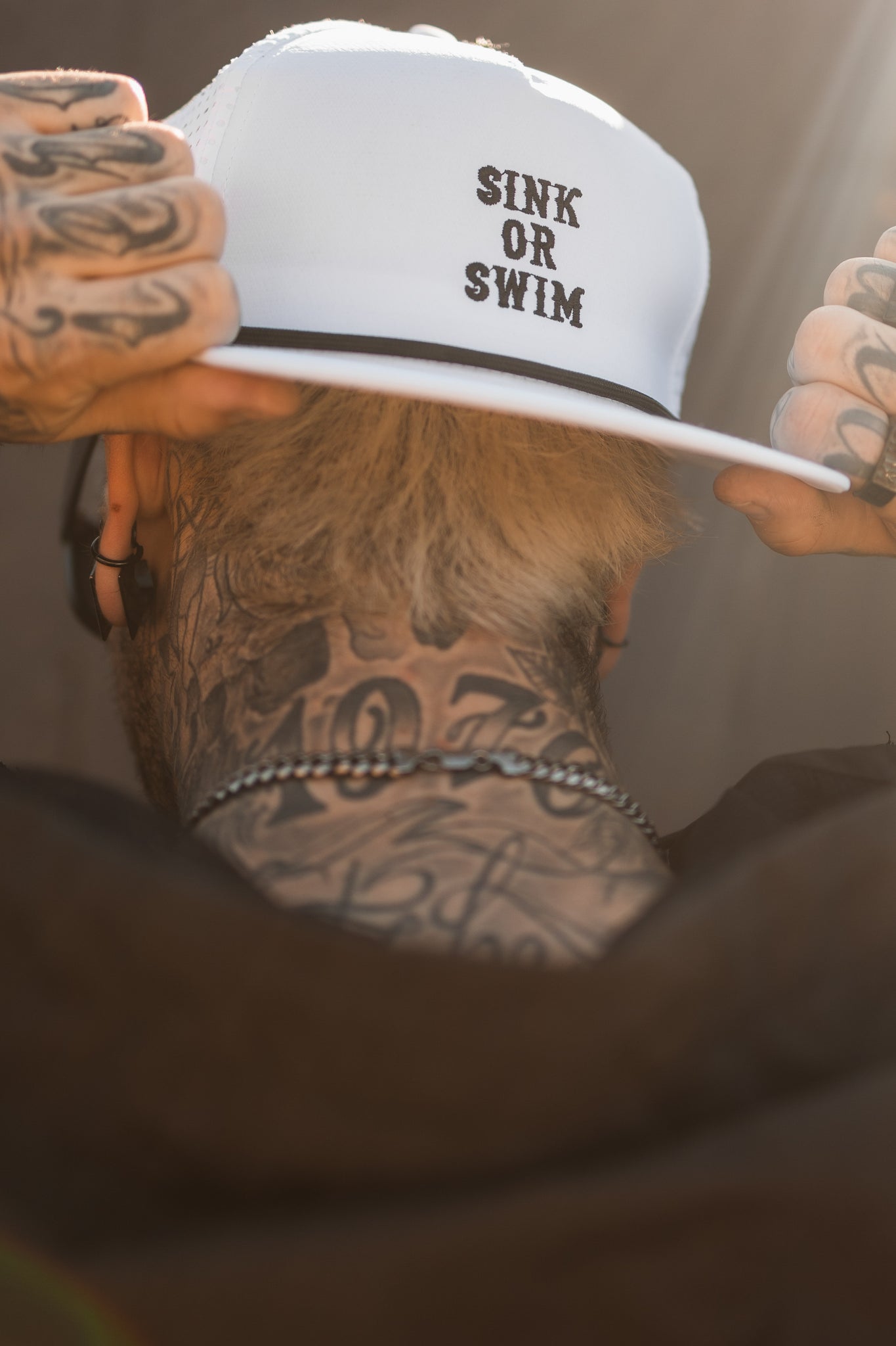A tattooed individual on their hands and neck adjusts a Relentless Betrayals Sink or Swim WHITEOUT Snapback. In the softly blurred background, sunlight creates a glow around their head.