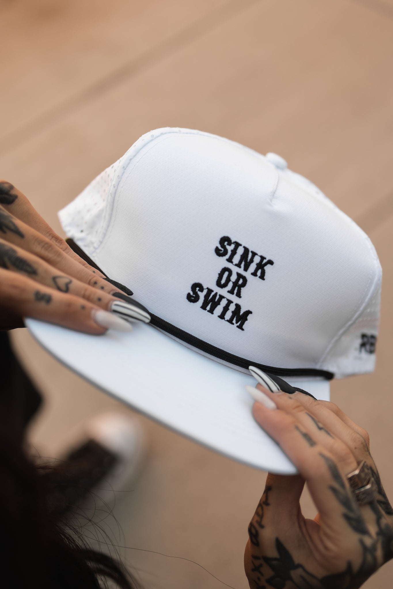 A person with tattooed hands and long nails holds the Sink or Swim WHITEOUT Snapback by Relentless Betrayal, a breathable white cap with SINK OR SWIM embroidered on the front. The background is blurred and neutral, emphasizing the caps stylish design.
