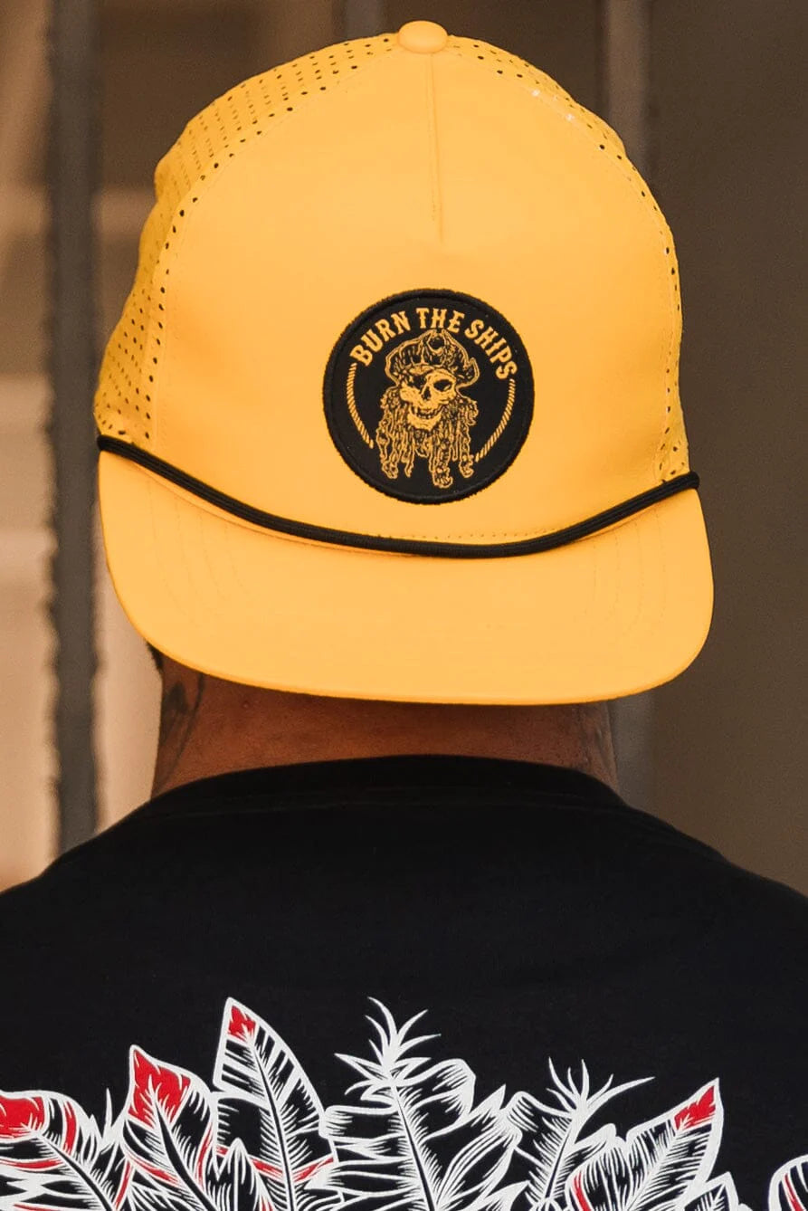 Wearing the Burn The Ships - Gold Snapback from Relentless Betrayal—a yellow hat with a pirate captain patch and BURN THE SHIPS text—complements their black shirt with colorful abstract designs in an indoor setting.