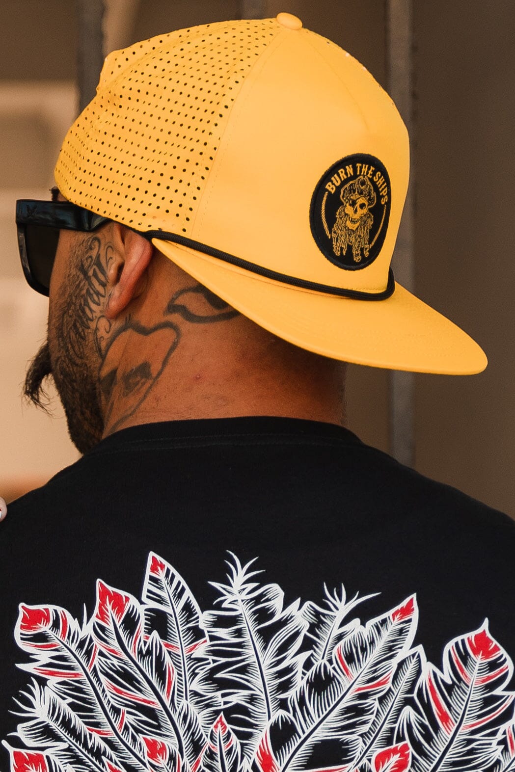 A person sports a Burn The Ships - Gold Snapback cap by Relentless Betrayal, featuring a pirate captain patch and logo. They pair it with sunglasses and a black T-shirt with white and red feathers, while their tattooed neck adds an edgy vibe.