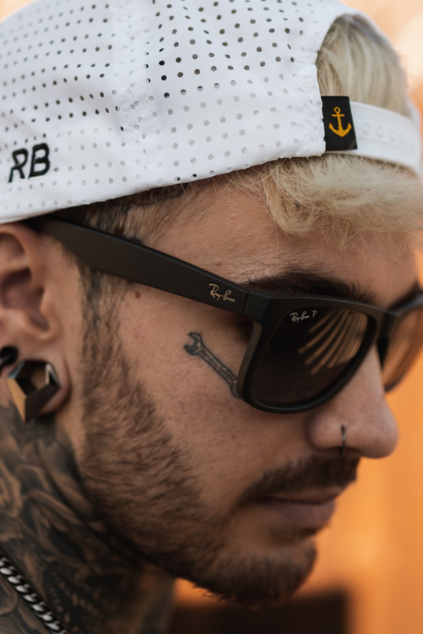 A bearded person wears sunglasses and flaunts a tattoo of a small bone near their shades while donning the Sink or Swim WHITEOUT Snapback by Relentless Betrayal, featuring an anchor logo and RB emblem.