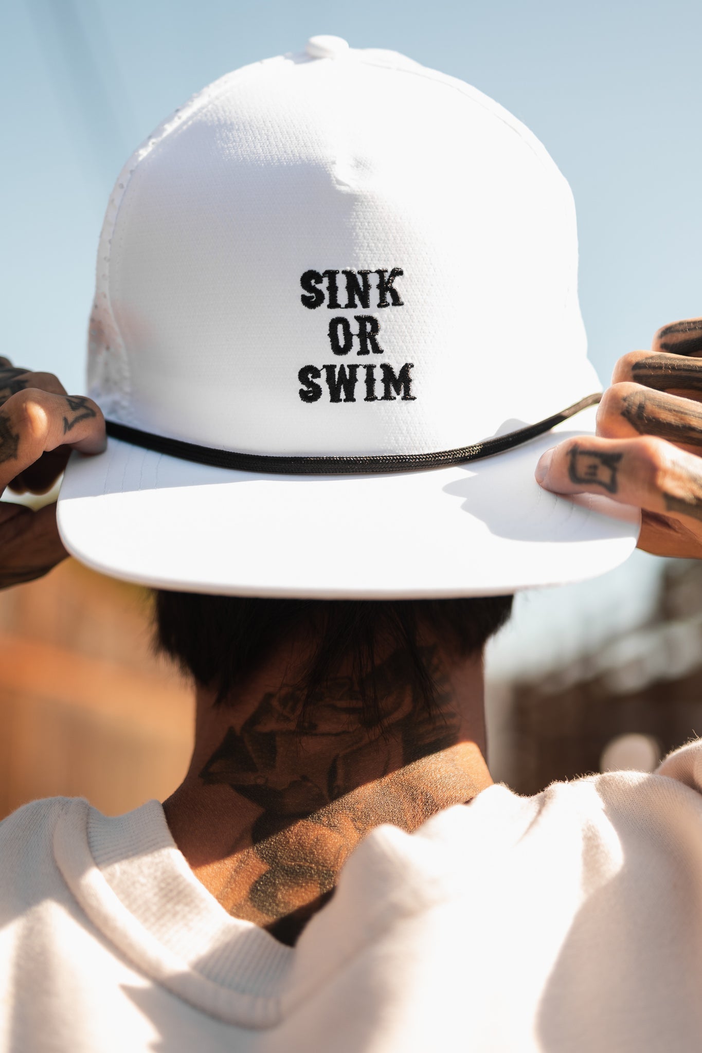 A tattooed individual dons a Relentless Betrayal Sink or Swim WHITEOUT Snapback, featuring black-embroidered SINK OR SWIM on the front. Theyre seen from behind against a blurred background.
