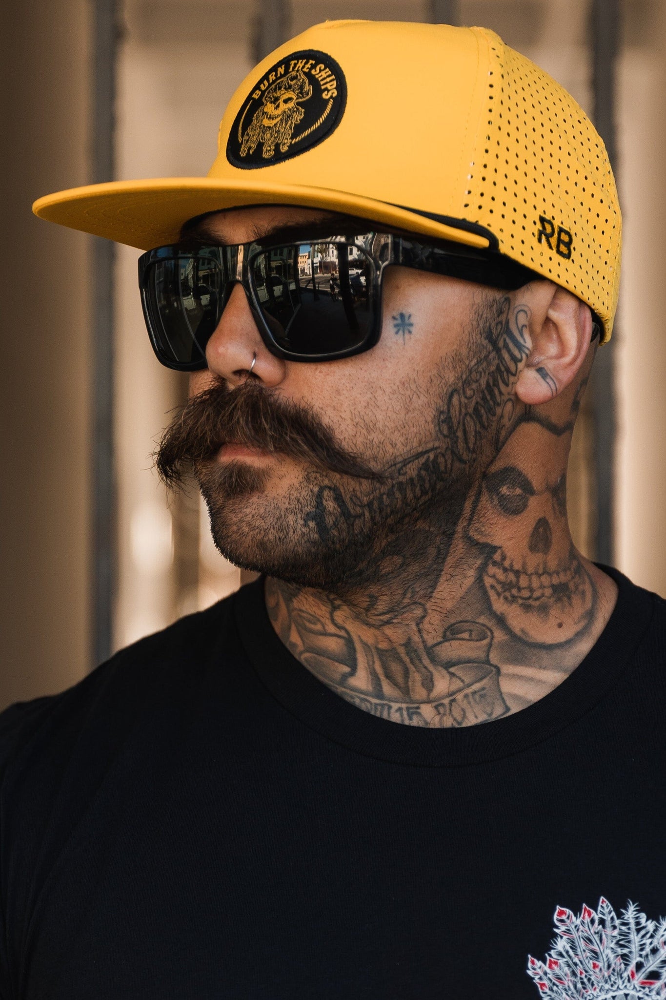 Sporting a large mustache and tattoos of skulls on their neck and arms, a person rocks the Burn The Ships - Gold Snapback by Relentless Betrayal, featuring a pirate captain patch. Their edgy look is completed with black sunglasses, a shirt, nose piercing, and earring.
