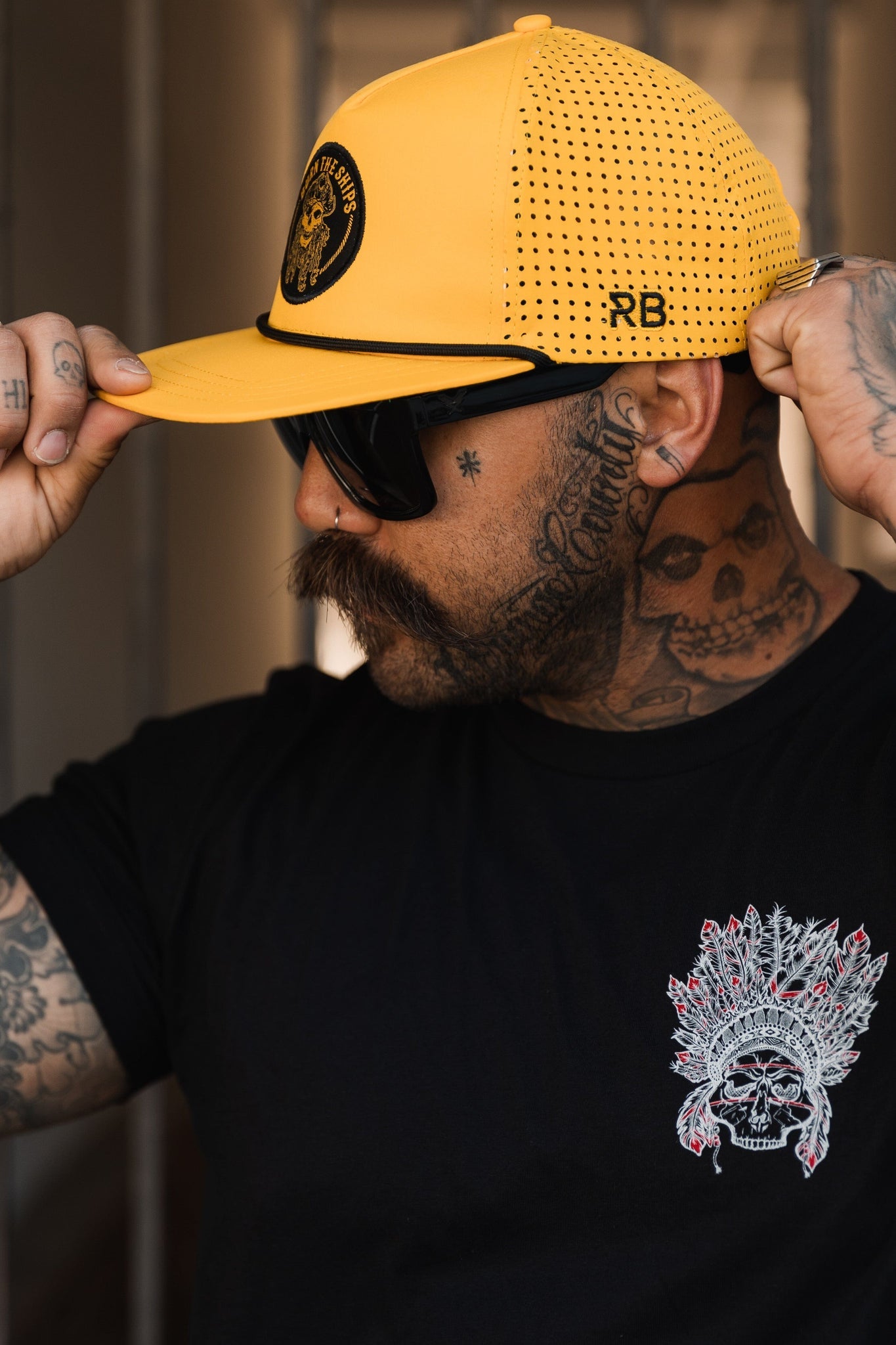 A tattooed person in a black t-shirt featuring a skull adjusts their Burn The Ships - Gold Snapback hat by Relentless Betrayal. They sport sunglasses, a large mustache, and tattoos on their neck and arms.