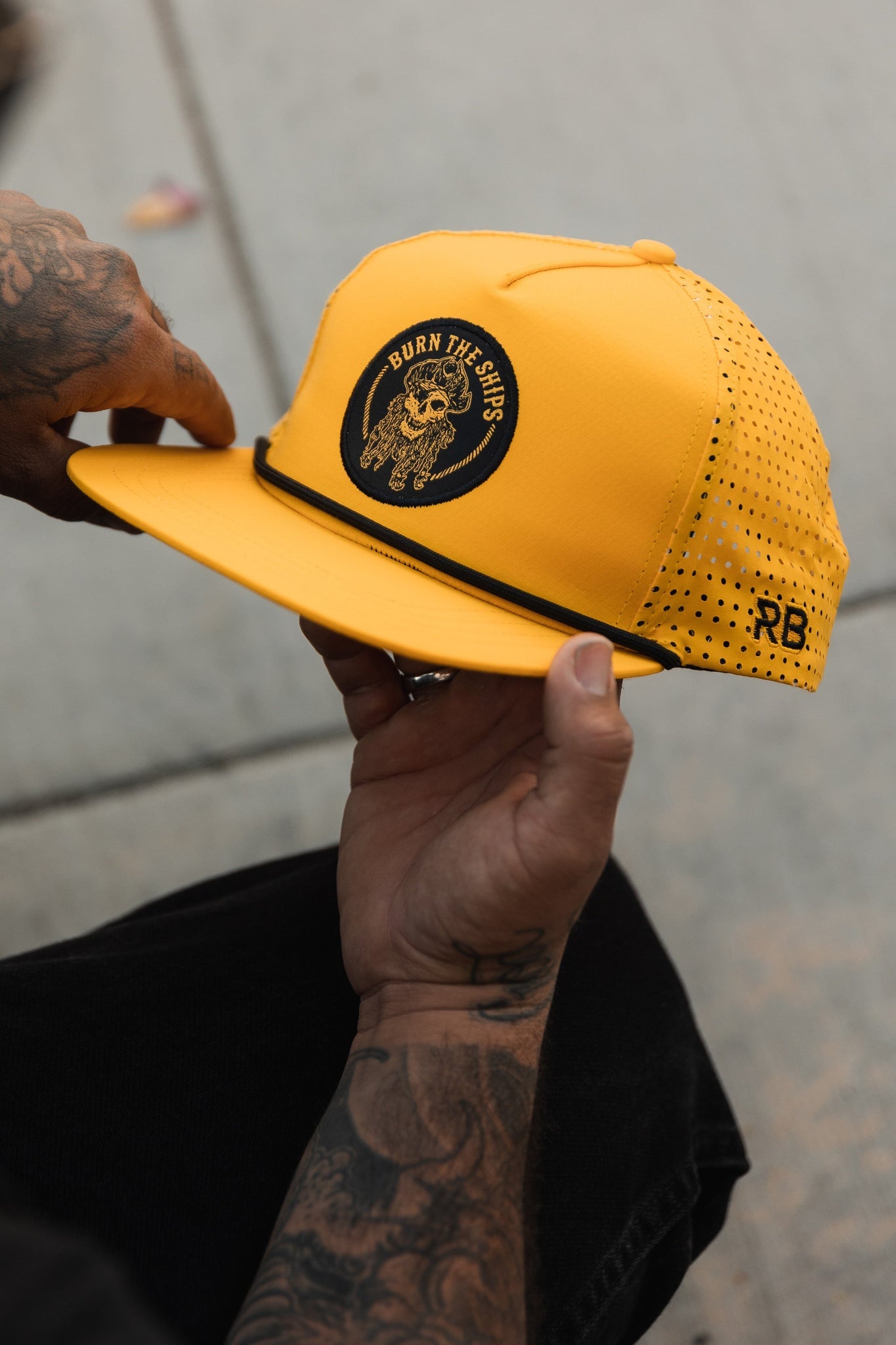 A tattooed individual holds the Burn The Ships - Gold Snapback by Relentless Betrayal, featuring a black embroidered Viking ship design and pirate captain patch. This water-resistant hat has perforated sides, RB initials, and is set on a concrete background.