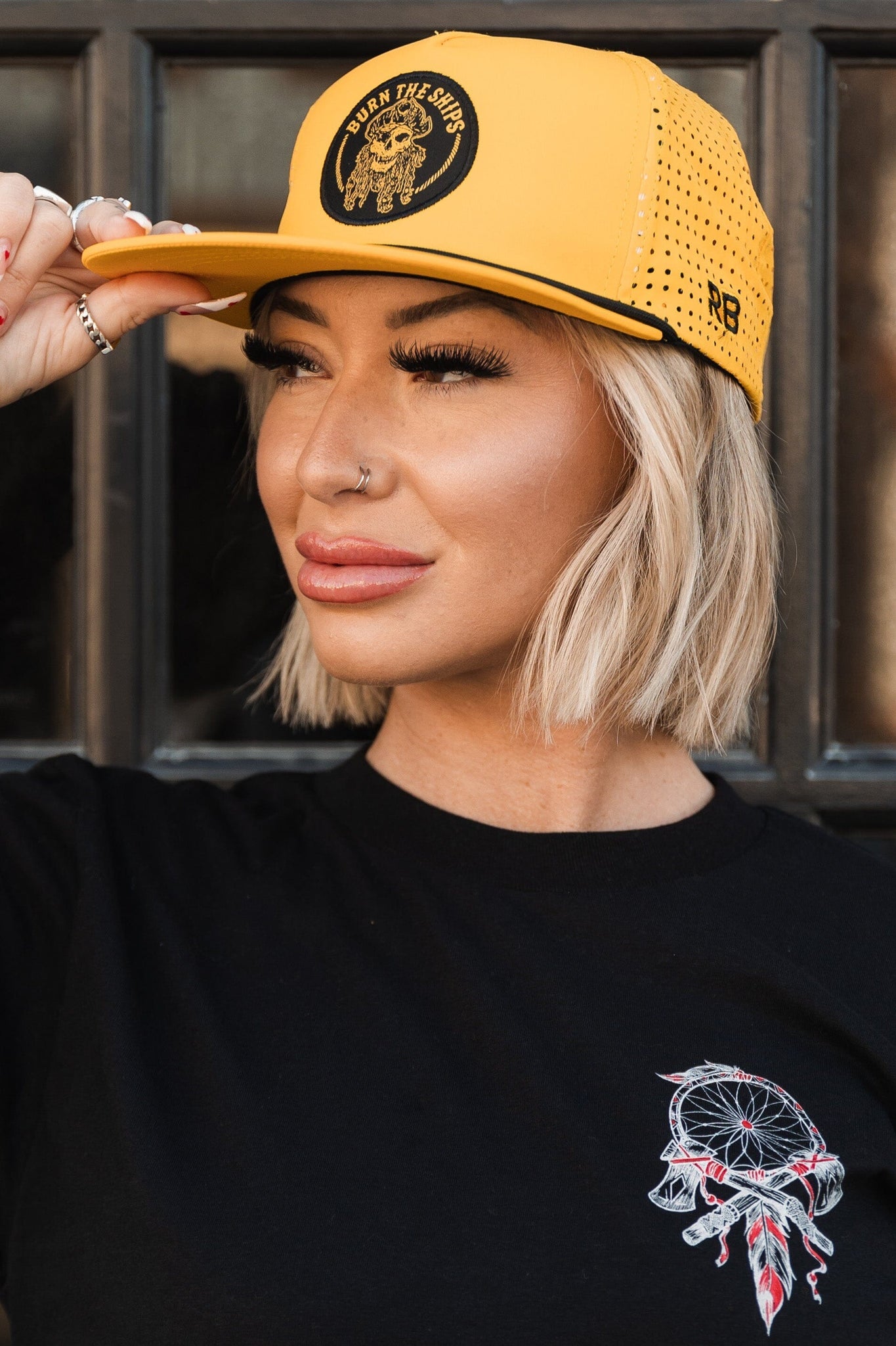 A woman in a black dreamcatcher t-shirt stands against a dark background, wearing the Burn The Ships - Gold Snapback by Relentless Betrayal. She has short blonde hair, a nose ring, and adjusts her cap with one hand.