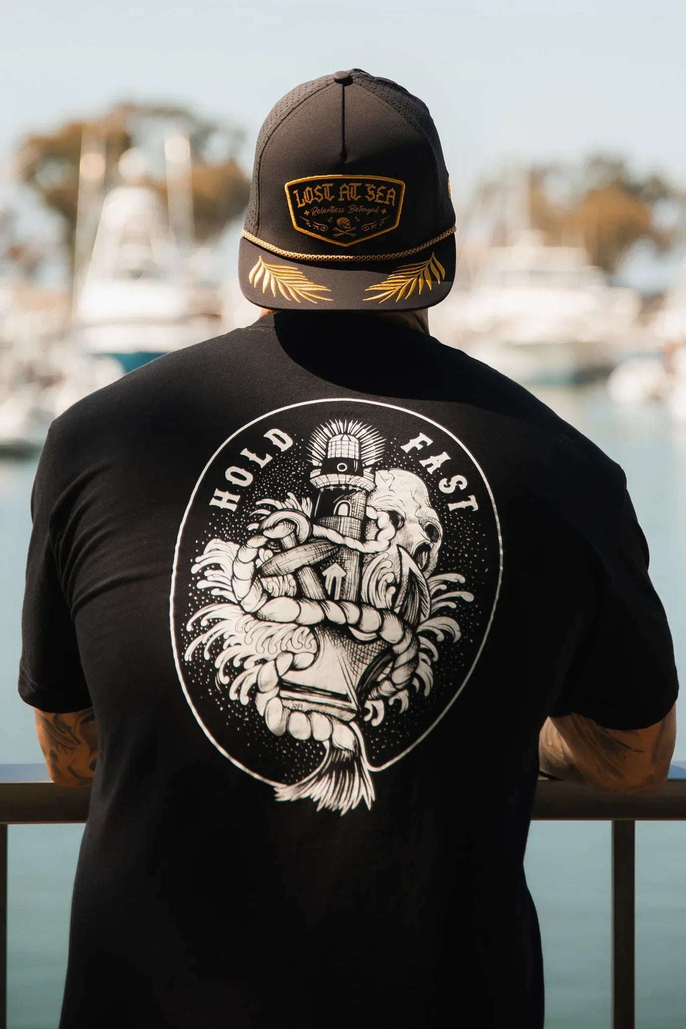 A person in a black Relentless Betrayal Lost at Sea cap and a Hold Fast T-Shirt—Black leans on a railing, gazing over a marina with blurred boats and water in the background.