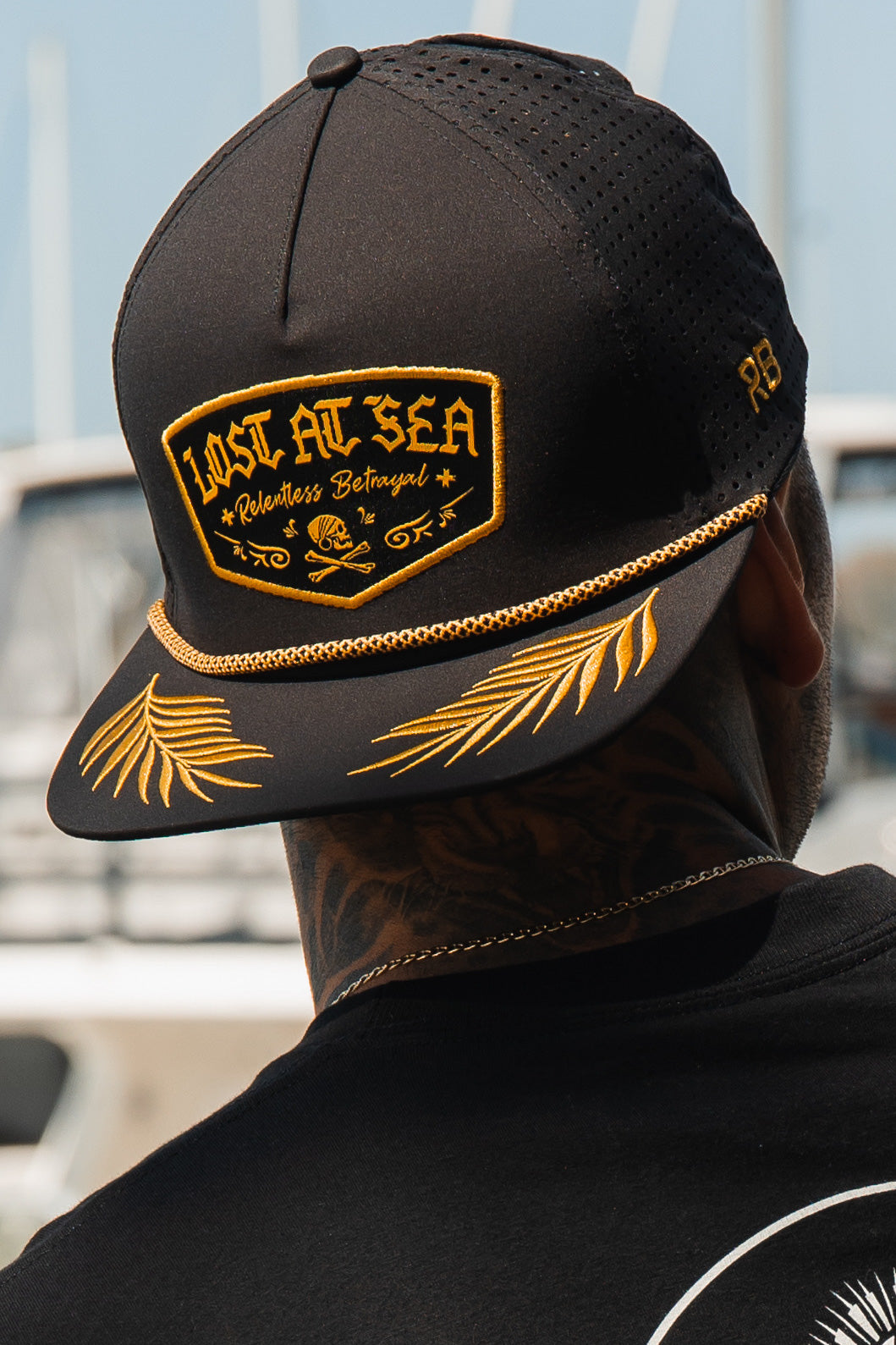 A man wears the Relentless Betrayals Lost at Sea Blackout Snapback, a black cap with gold embroidery stating LOST AT SEA above a mini skull and crossbones. This stylish, water-resistant hat has a snapback closure, set against blurred boats on shimmering waters.