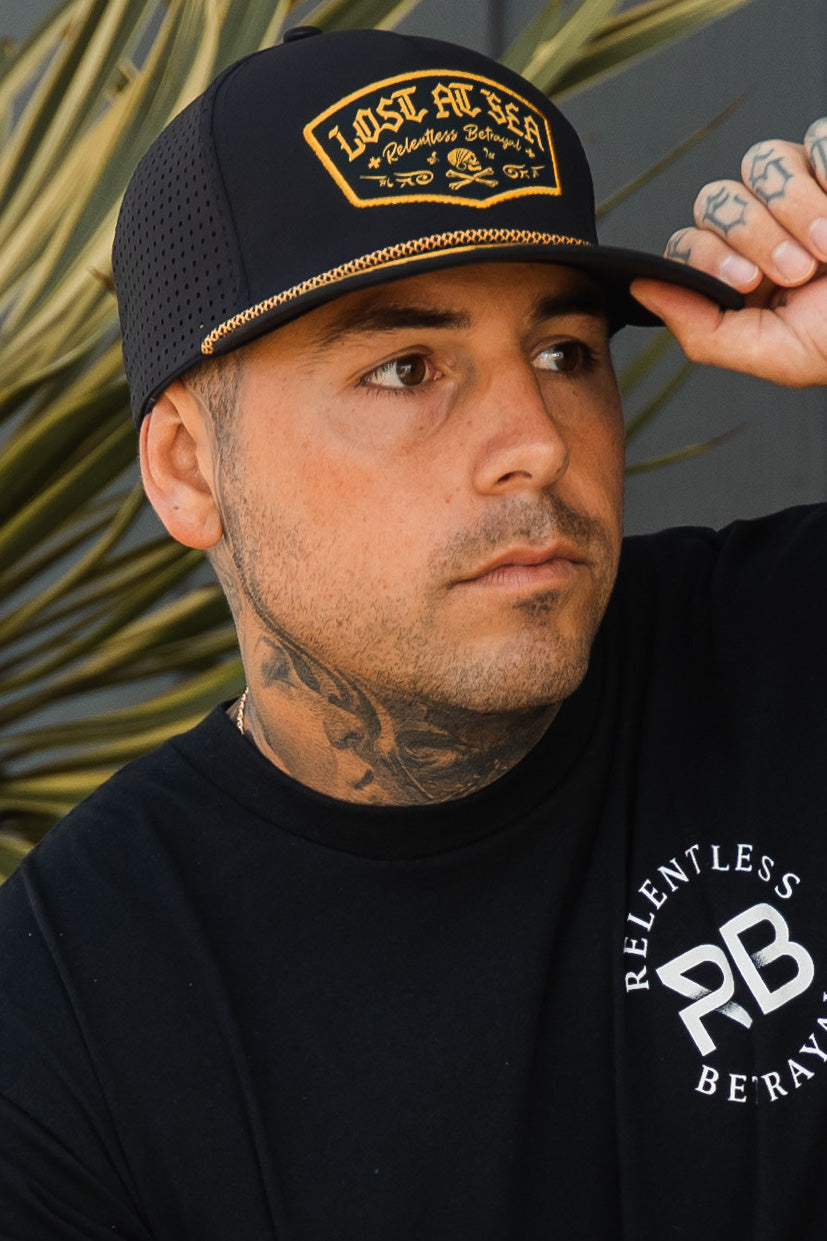 Wearing a Lost at Sea Blackout Snapback and a Relentless Betrayal T-shirt, tattoos on their neck and hand show as they touch the caps brim. A blurred plant in the background adds to the mysterious vibe.