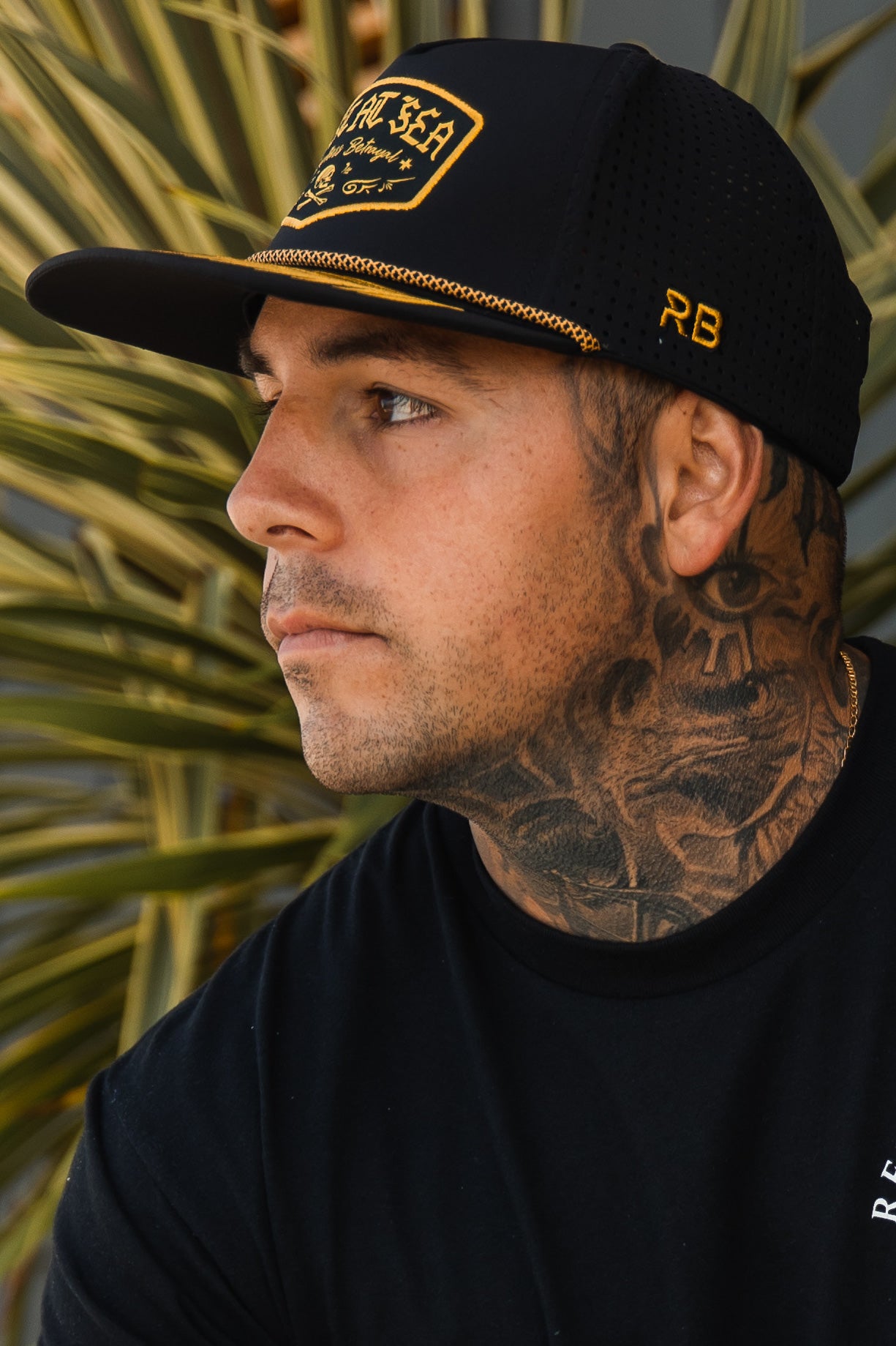 A person with a tattooed neck wears the Lost at Sea Blackout Snapback by Relentless Betrayal, black with yellow lettering. Looking aside in a black T-shirt, they stand against green palm leaves as the water-resistant hat is secured by its snapback enclosure.