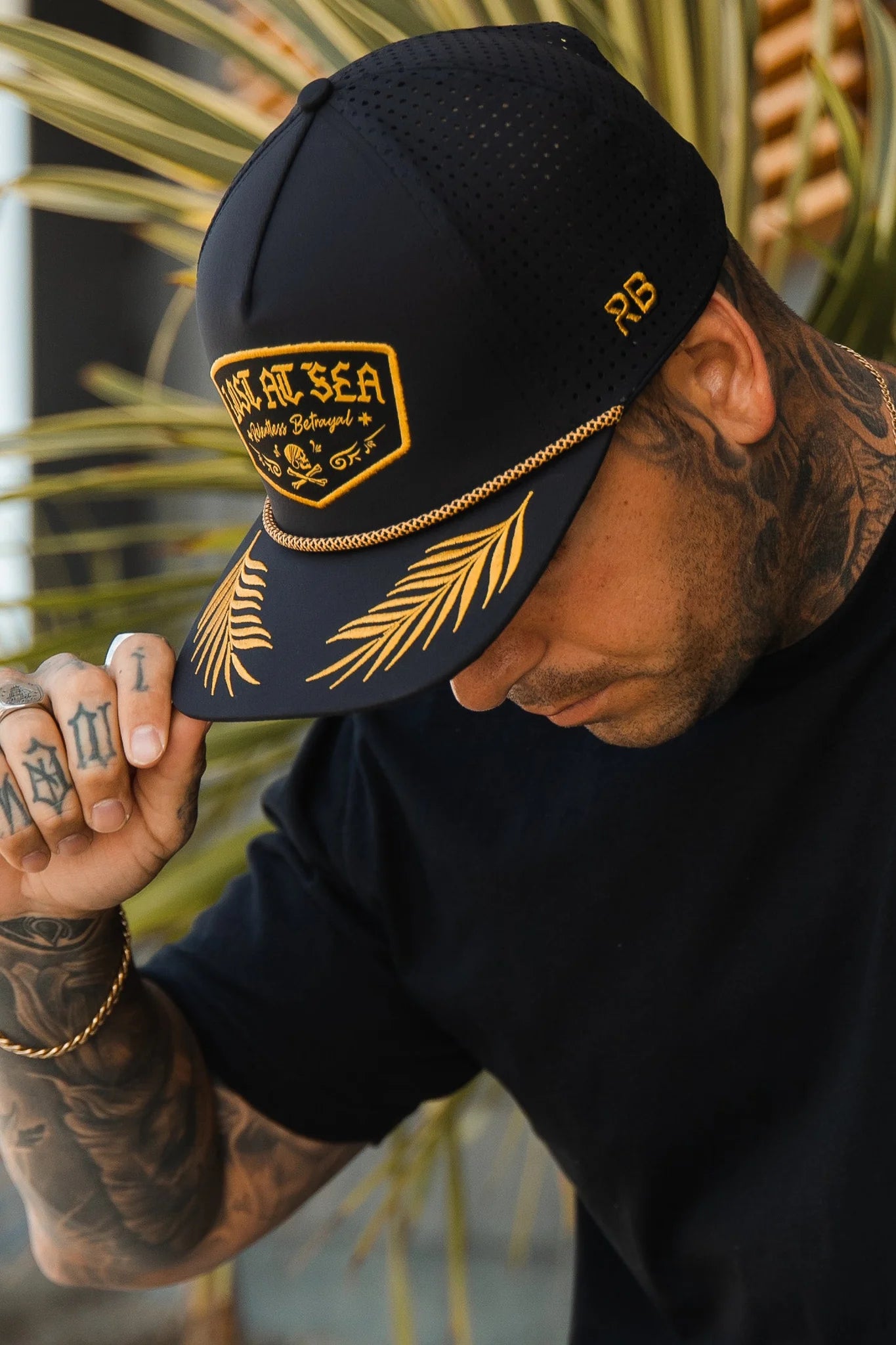 A man in a black Lost at Sea Blackout Snapback by Relentless Betrayal adjusts the caps snapback enclosure with his hand. Hes wearing a black t-shirt, showcasing tattoos on his arms and hand, with a blurred plant in the background.