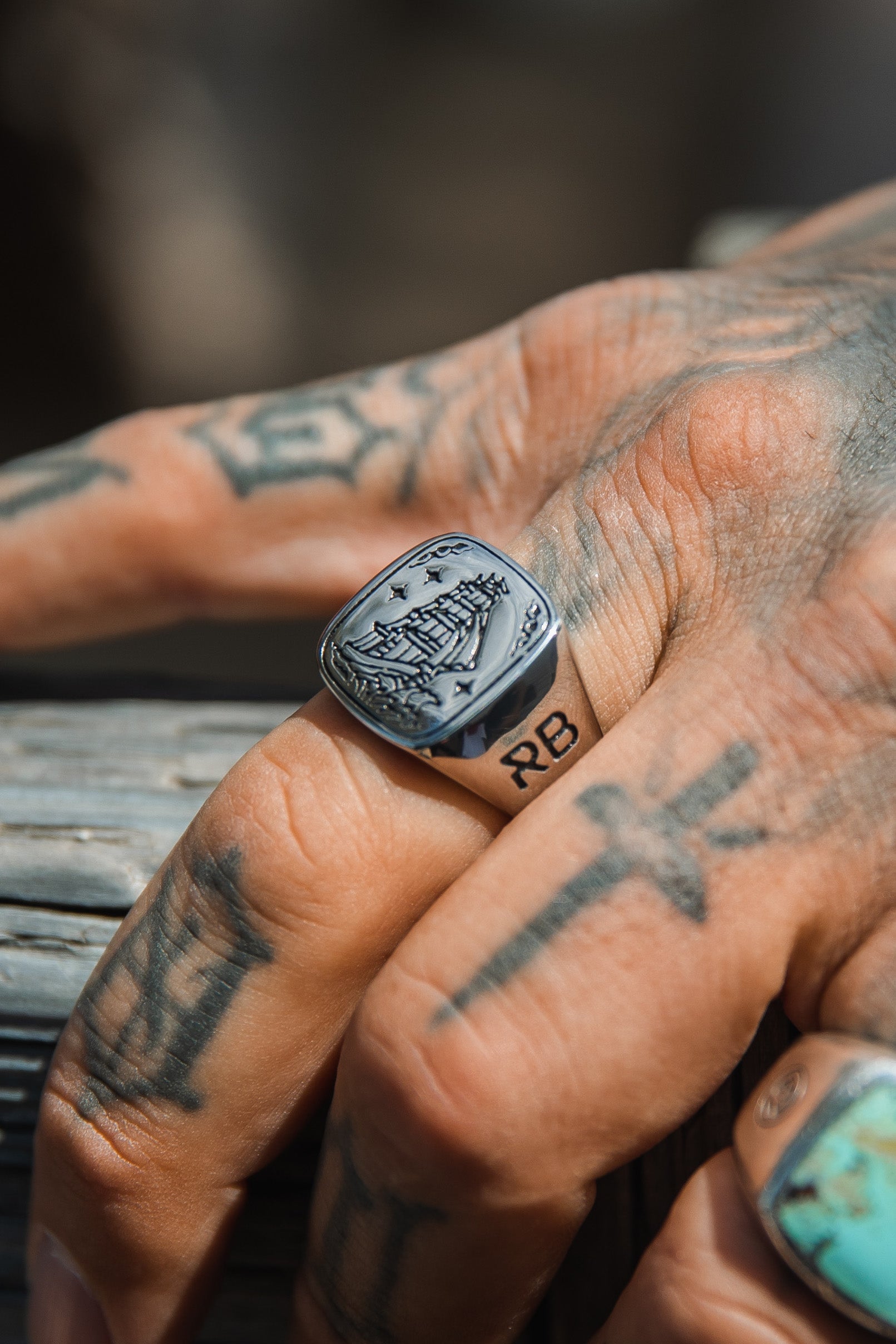 A tattooed hand showcases the Relentless Betrayals Lost At Sea Ring with a ship design in stainless steel. The index finger, marked with an RB tattoo amidst abstract designs, rests on wood.