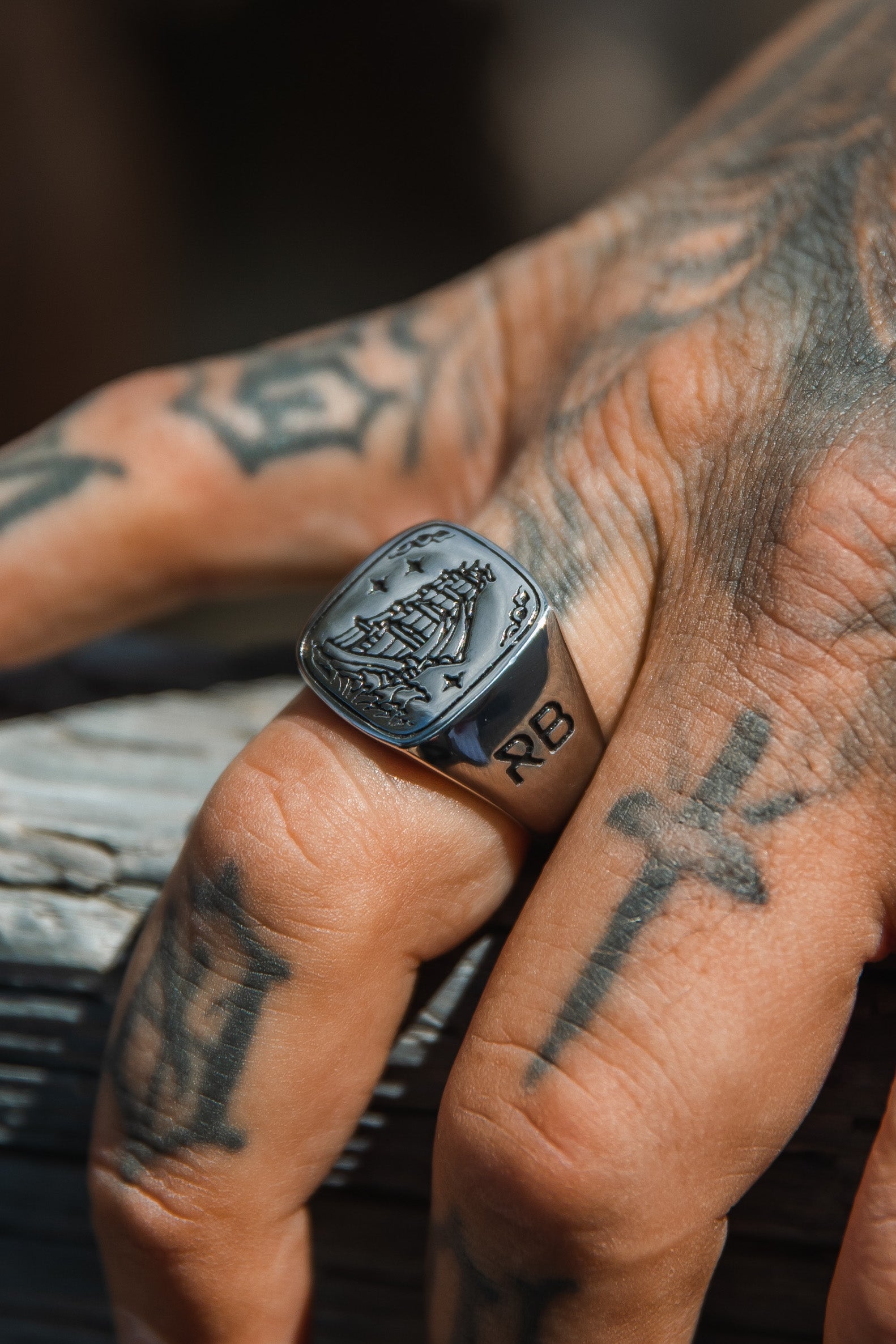 Close-up of a tattooed hand on wood, wearing Relentless Betrayals Lost At Sea Ring, intricately engraved with a ship. Sunlit tattoos—a cross and letters—enhance the rugged nautical theme, while its stainless steel design completes the look.