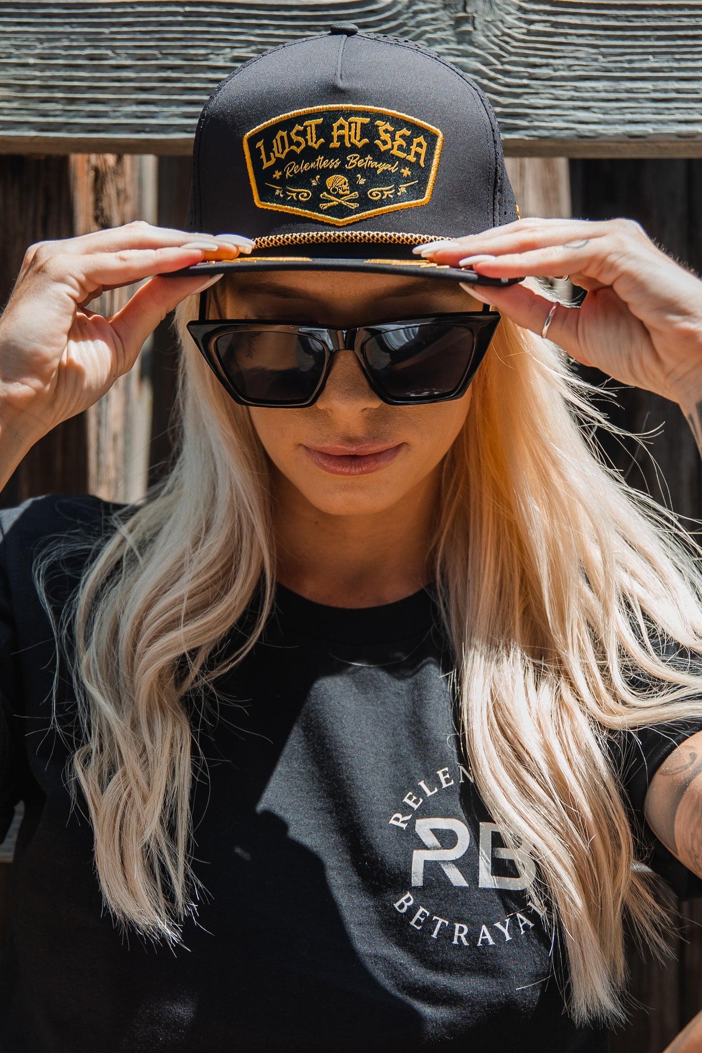 A long-haired blonde wears a Lost at Sea Blackout Snapback, large sunglasses, and a black Relentless Betrayal T-shirt. The wood paneled backdrop adds an inviting allure to the scene.