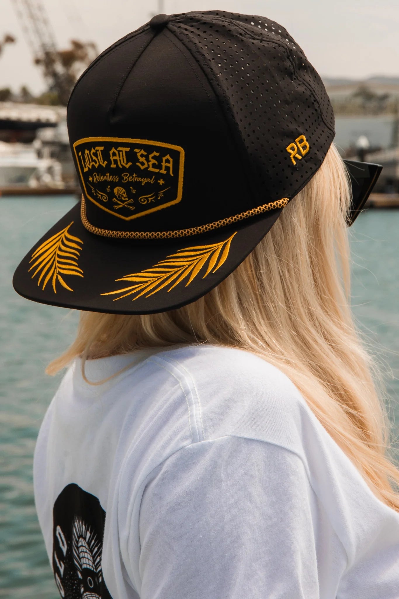 A person with long blonde hair wears a Relentless Betrayal Lost at Sea Blackout Snapback with black fabric and yellow embroidery by the marina. The snapback adds style to their look, complemented by a white shirt featuring a black and white design on the back.