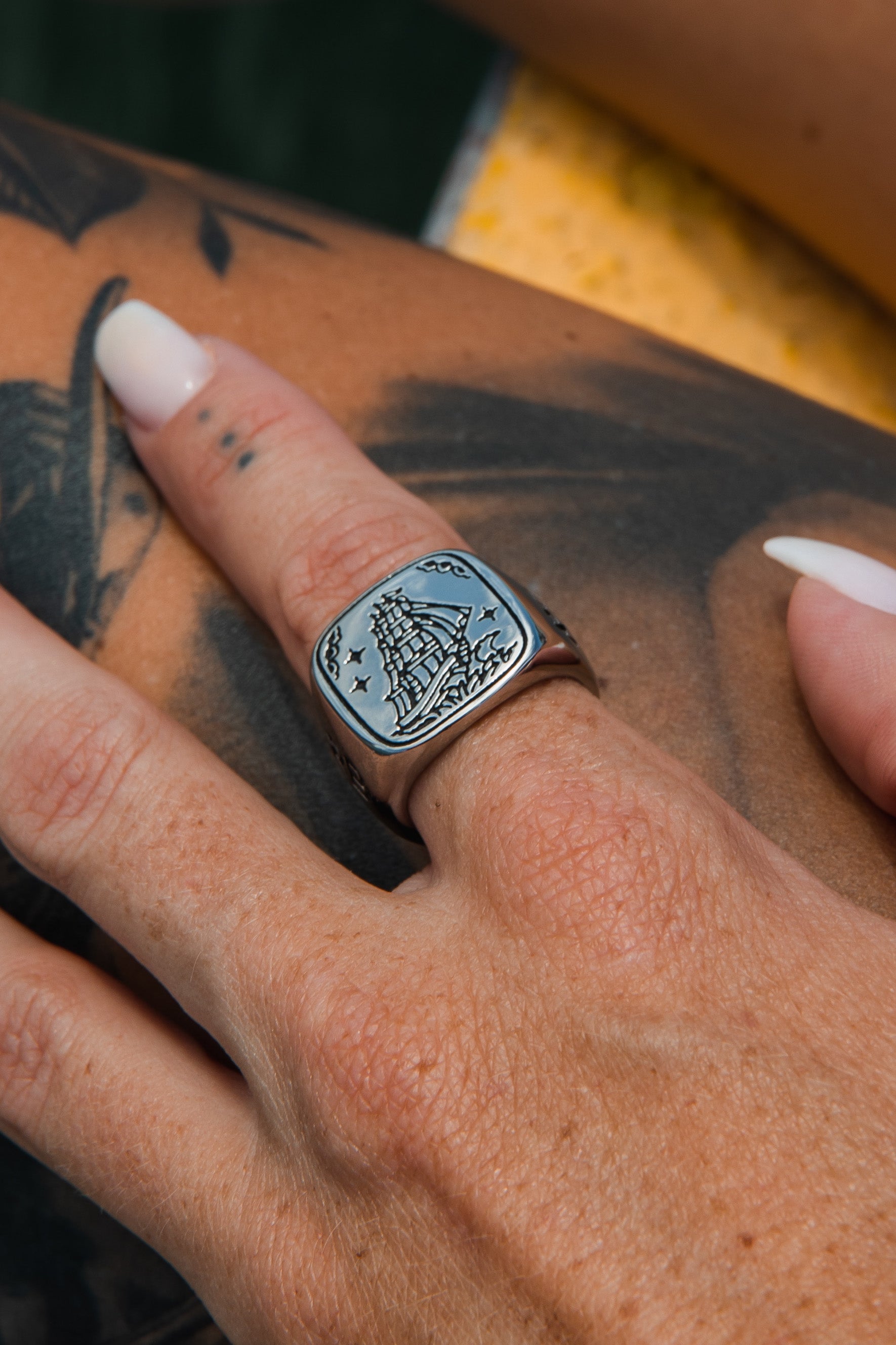 A hand with a tattoo displays Relentless Betrayals handcrafted Lost At Sea Ring, a silver piece with an engraved ship design. The manicured nails are white, and the blurred background combines green and yellow hues, creating an atmosphere of stainless steel elegance.