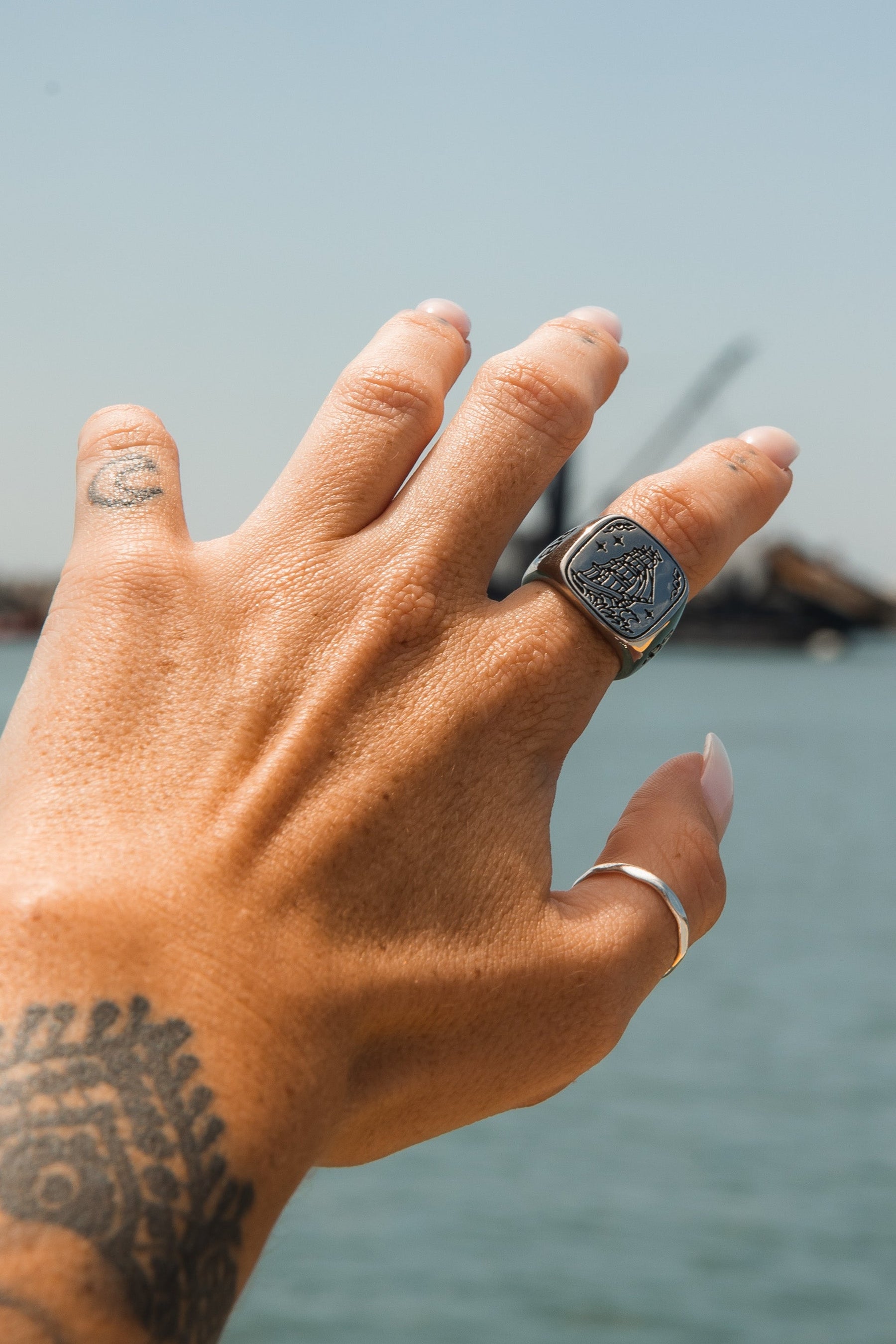 A tattooed hand with polished nails stands against a blurred backdrop of water and a crane, showcasing the handcrafted Relentless Betrayals Lost At Sea Ring with intricate design, paired elegantly with a slender stainless steel band on another finger.