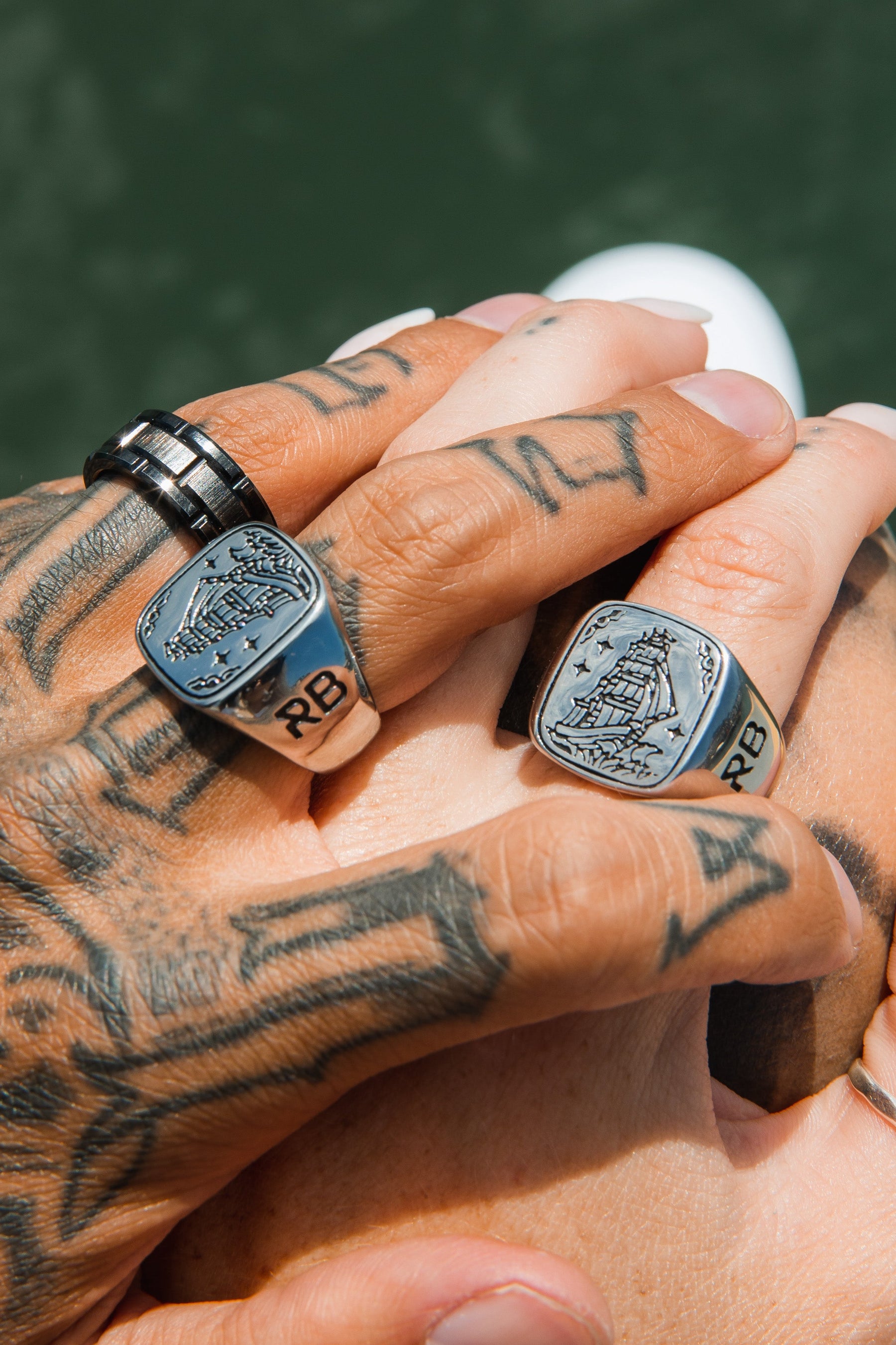 Tattooed hands with silver rings take center stage, featuring the exquisite Relentless Betrayals Lost At Sea Ring adorned with a ship and initials RB. Each handcrafted piece stands out against a blurred background, highlighting their intricate designs.
