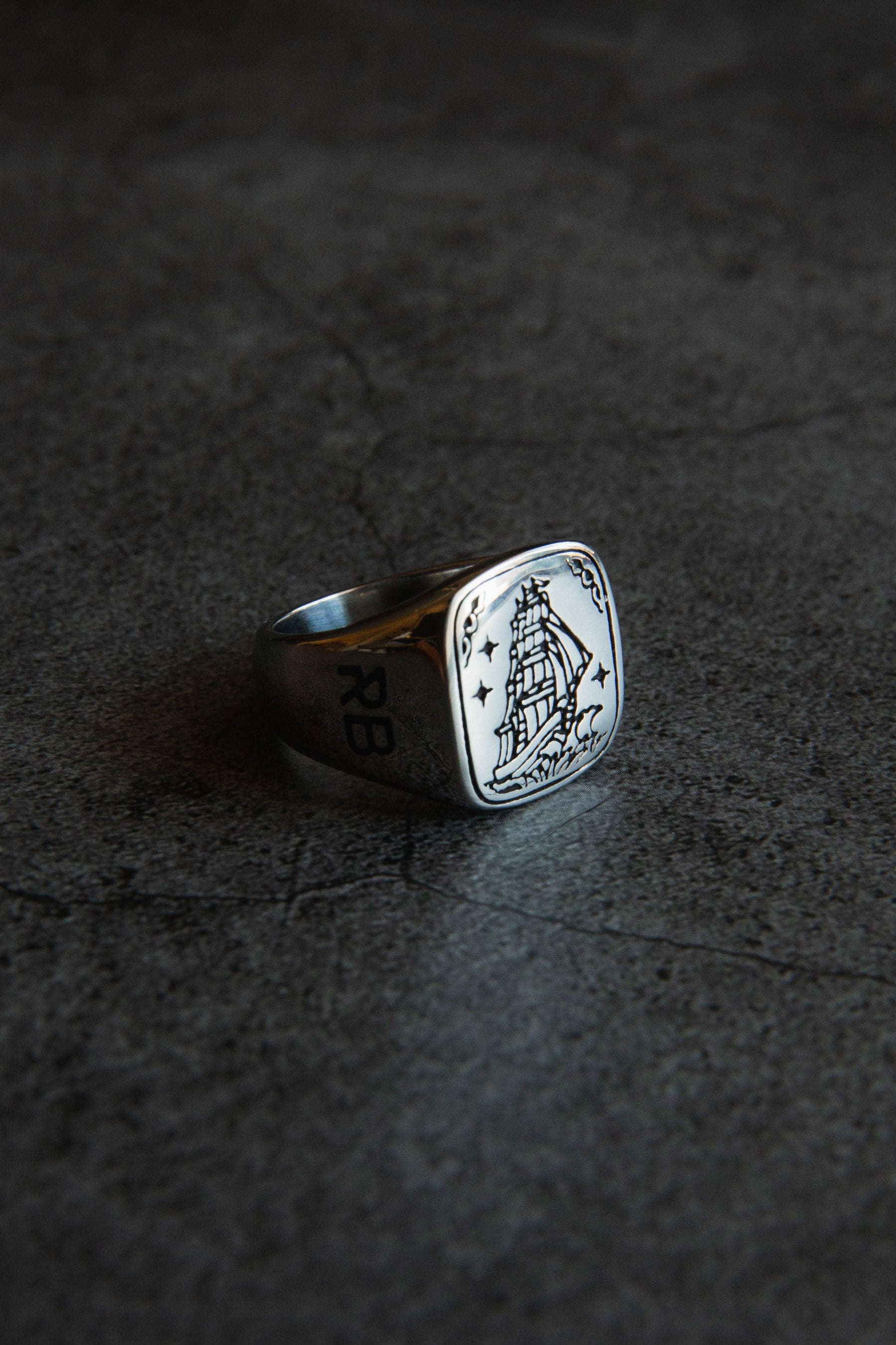 Introducing the Lost At Sea Ring by Relentless Betrayal: a stainless steel masterpiece with an engraved sailing ship and stars on its square face, set on a textured dark surface. The band features RB, showcasing handcrafted precision and artistry.