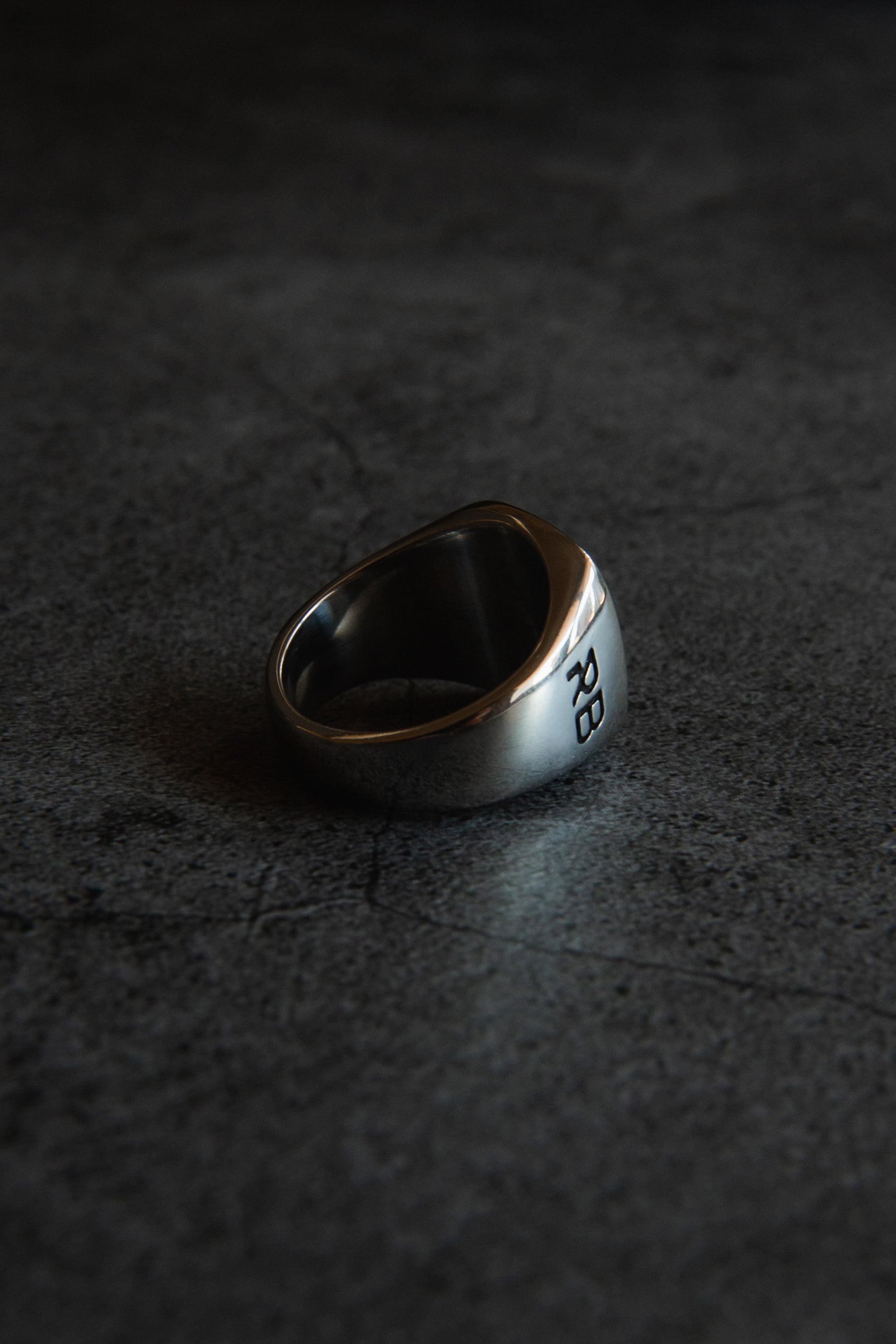 The Lost At Sea Ring from Relentless Betrayal, a handcrafted silver piece with RB initials engraved, sits on a dark, textured surface under dramatic lighting.