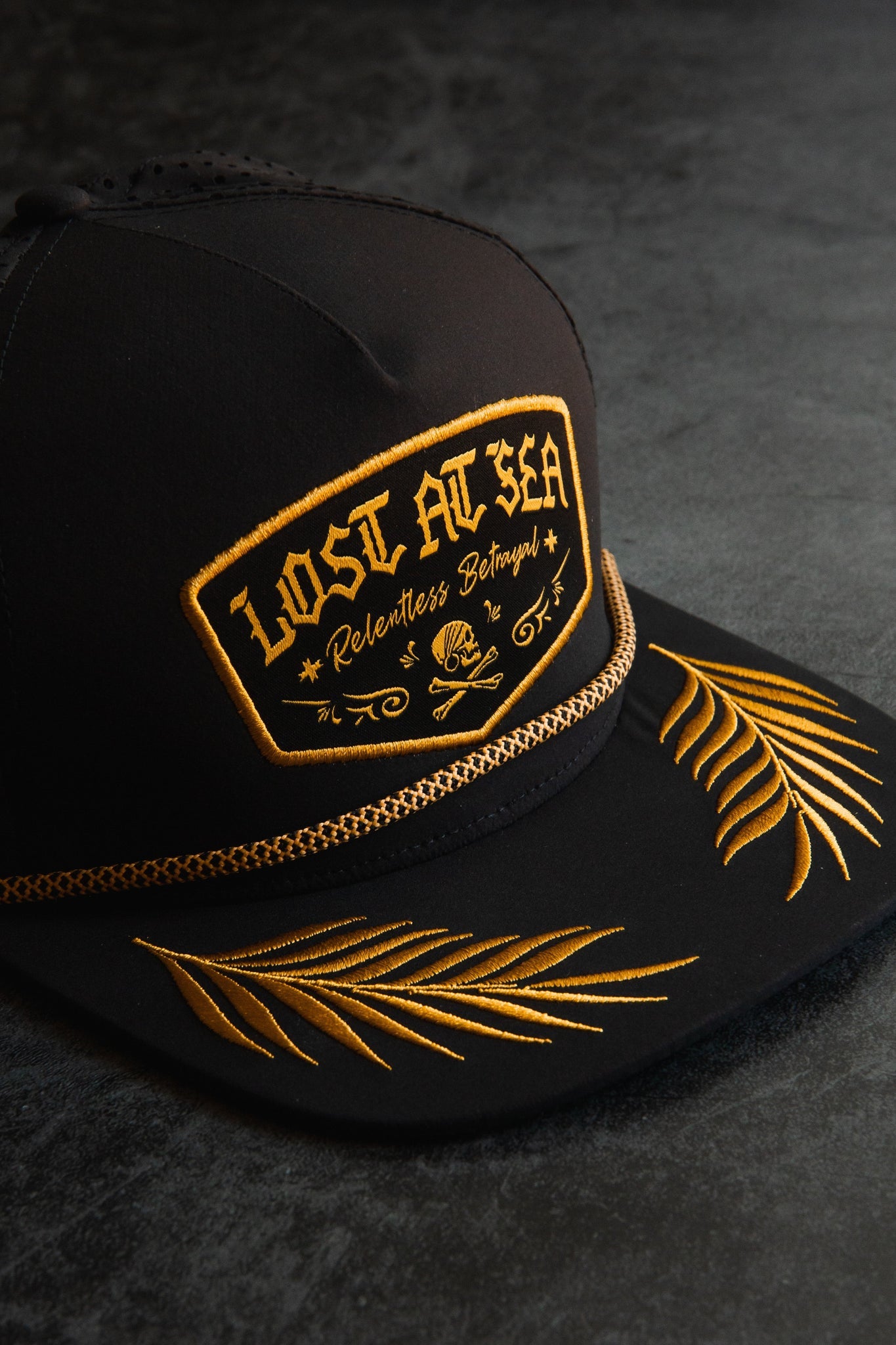 A Lost at Sea Blackout Snapback by Relentless Betrayal lies on a dark surface. It boasts gold embroidery and a patch with LOST AT SEA - Relentless Betrayal alongside a skull and crossed anchors. The water-resistant hat features stitched palm frond designs on the brim.