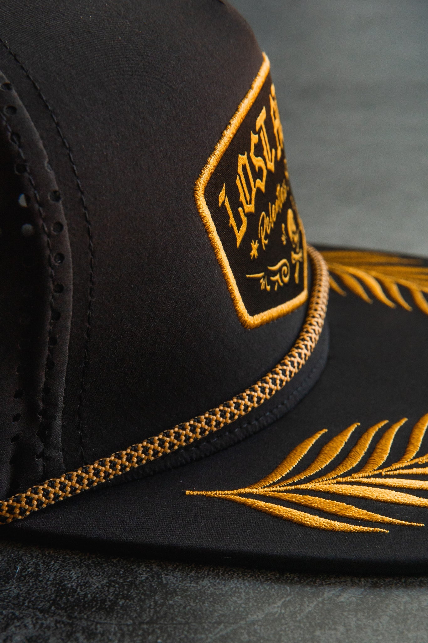 Close-up of the Relentless Betrayal Lost at Sea Blackout Snapback, showcasing a black design with an embroidered patch featuring gold lettering and designs. The hat has gold stitching on the brim leaves and a decorative front chain against a smooth, dark background.