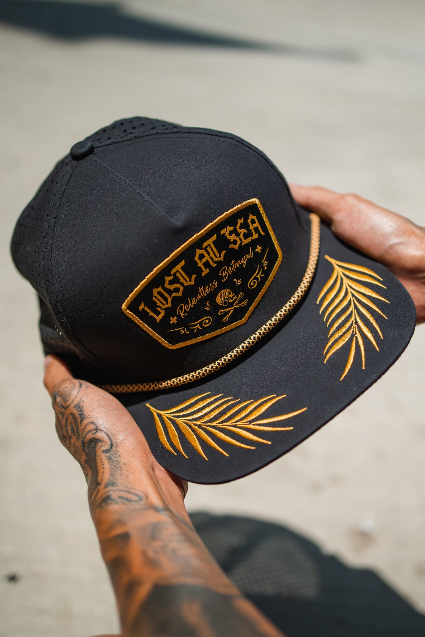 A tattooed person holds the stylish Lost at Sea Blackout Snapback by Relentless Betrayal, featuring gold embroidery and nautical designs on the brim, topped with a decorative gold chain. This water-resistant cap combines functionality with fashion.