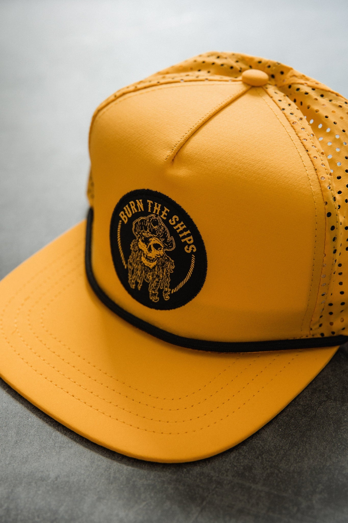 The Burn The Ships - Gold Snapback by Relentless Betrayal features a black pirate captain patch with a skull and Burn The Ships text on a yellow cap. It has a snapback closure, perforated back, and sleek black line above the visor, all resting on a gray surface.