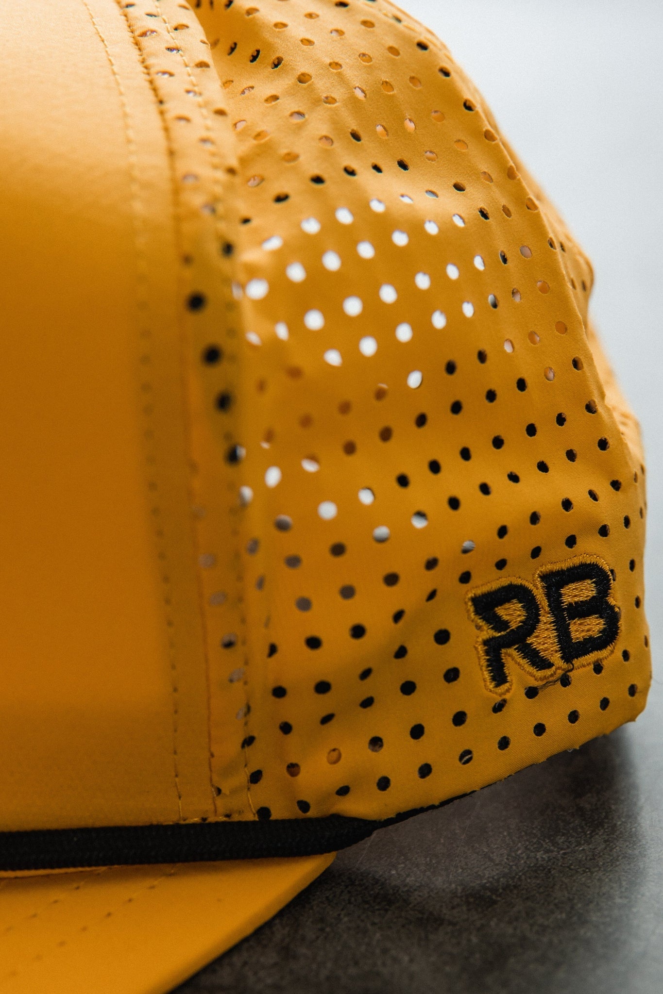 Close-up of the Burn The Ships - Gold Snapback by Relentless Betrayal, showcasing its yellow color with black perforated dots and RB embroidery. The snapback enclosure and textured details stand out against a blurred background.