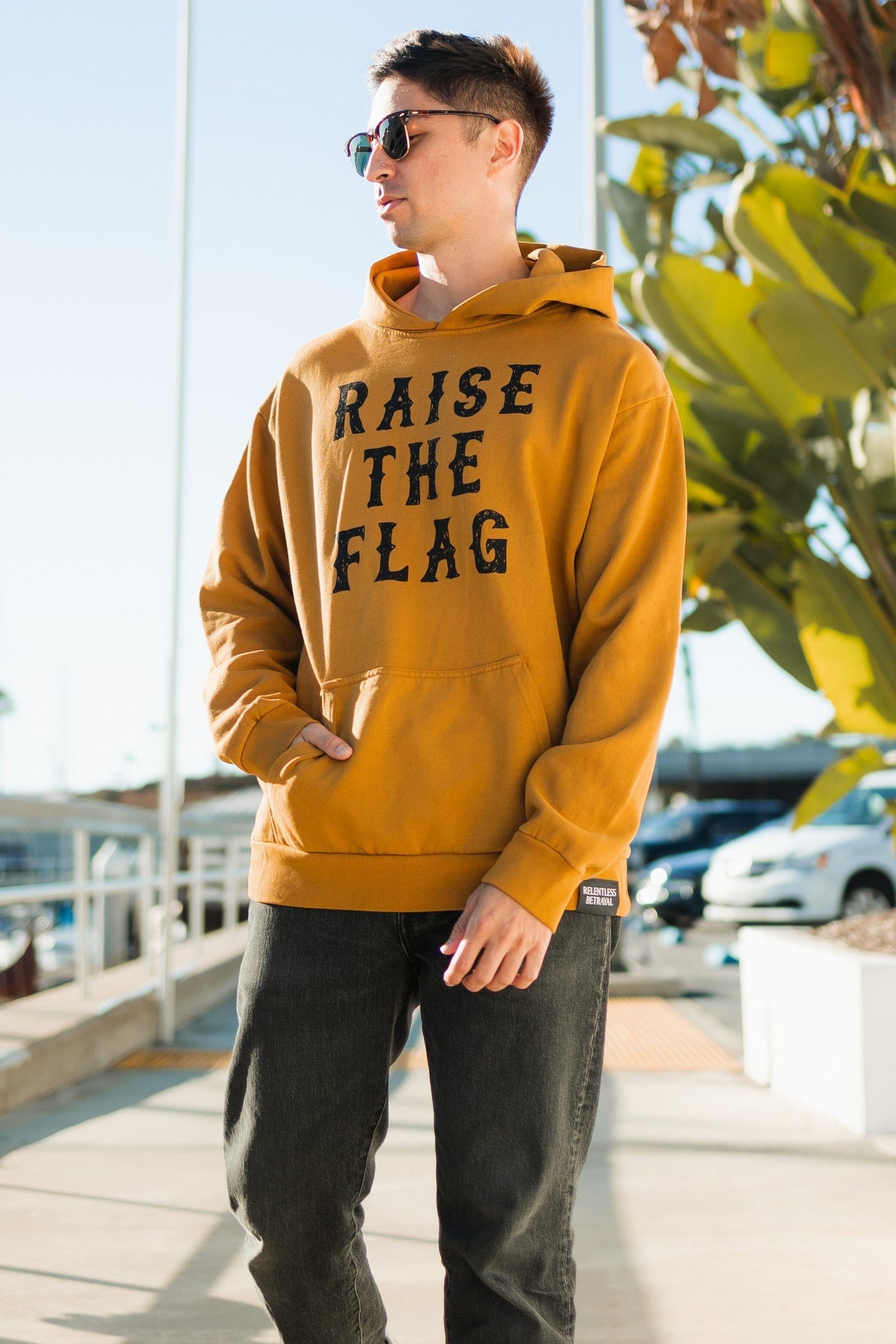 A person in a Raise the Flag Premium Toast Hoodie by Relentless Betrayal stands on a sunny sidewalk wearing sunglasses, with large green leaves and parked cars in the background, enhancing the relaxed vibe.