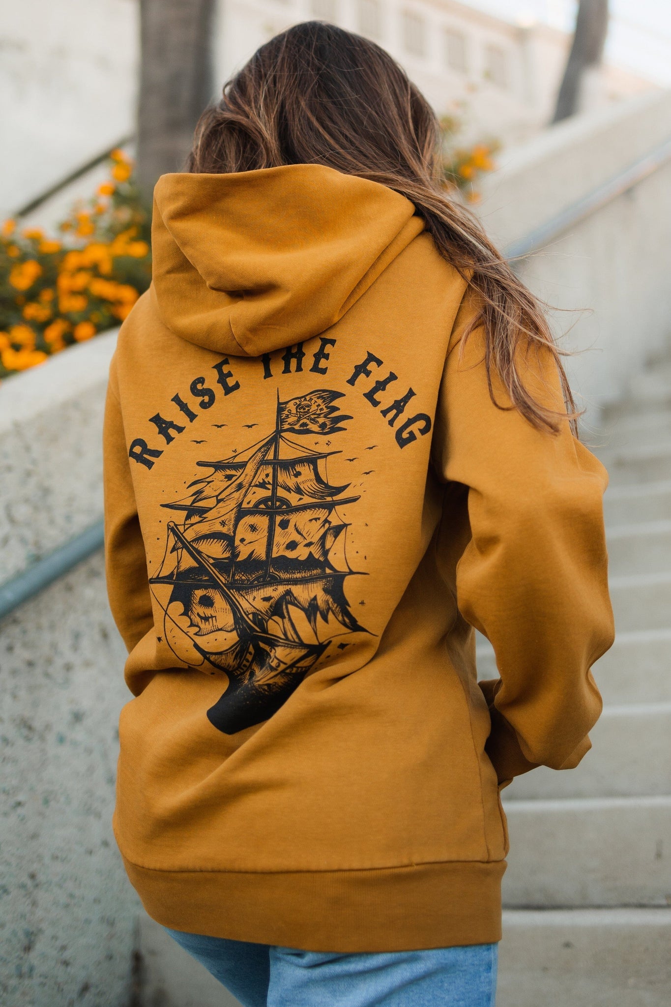 A person with long hair stands on outdoor steps next to an orange flower bed, wearing the unisex Relentless Betrayal Raise the Flag Premium Toast Hoodie in mustard with a pirate ship illustration and Raise the Flag text on the back.