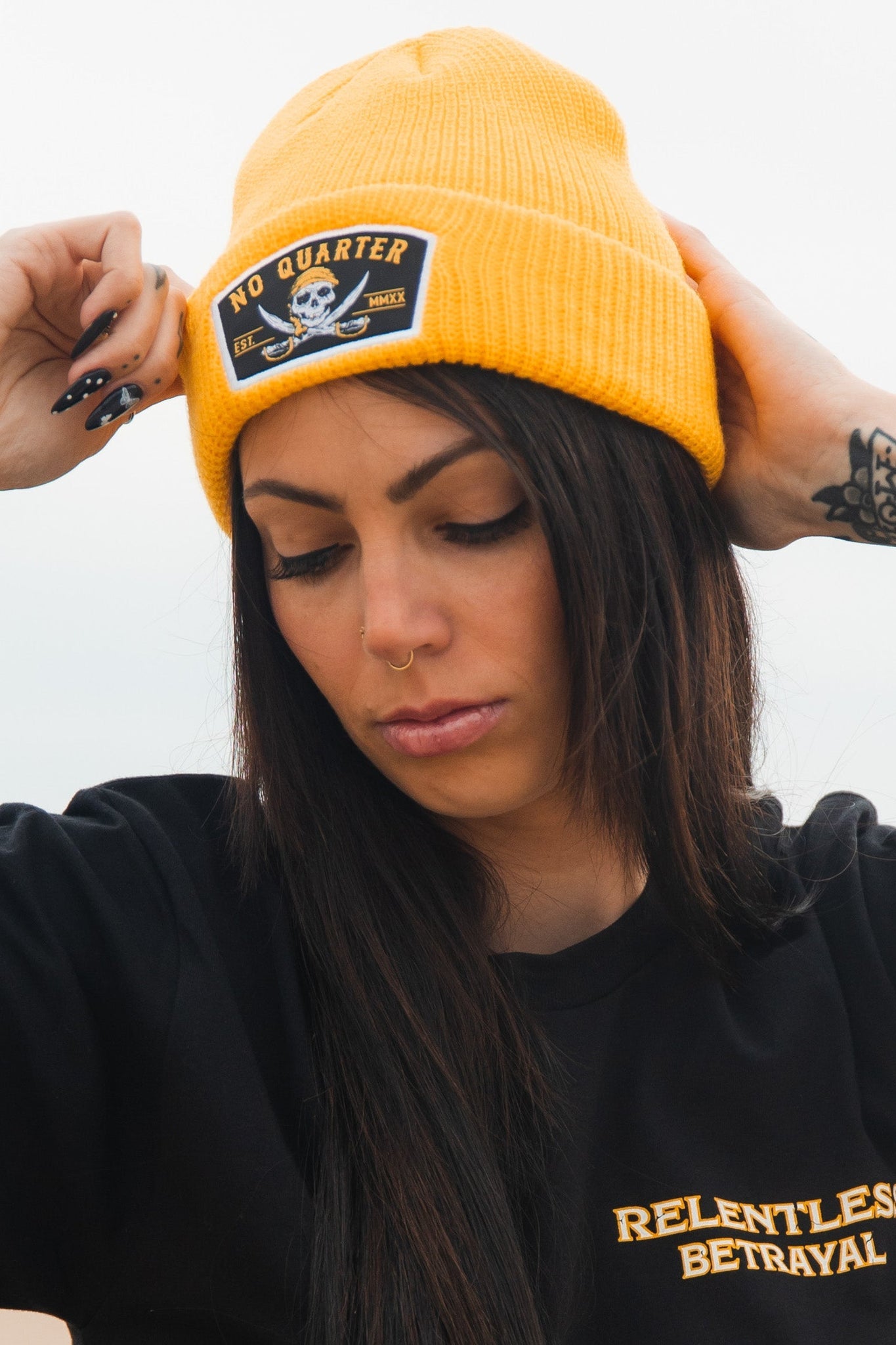 A person adjusts their No Quarter Beanie with both hands, showcasing long dark hair and a nose ring. Theyre wearing a black Relentless Betrayal shirt. The blurred background evokes the mystique of the classic skull and crossbones symbol.